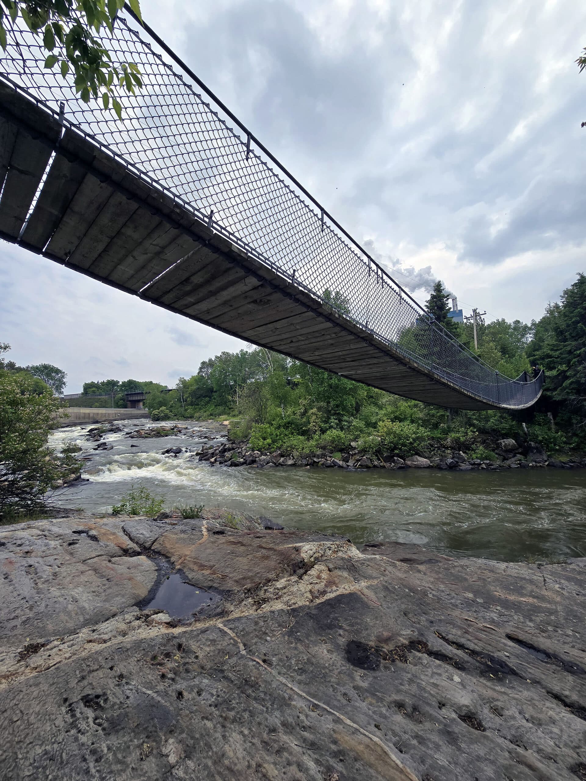 The Roy Wilson suspension bridge as seen from below and the side, with a small waterfall in the background.