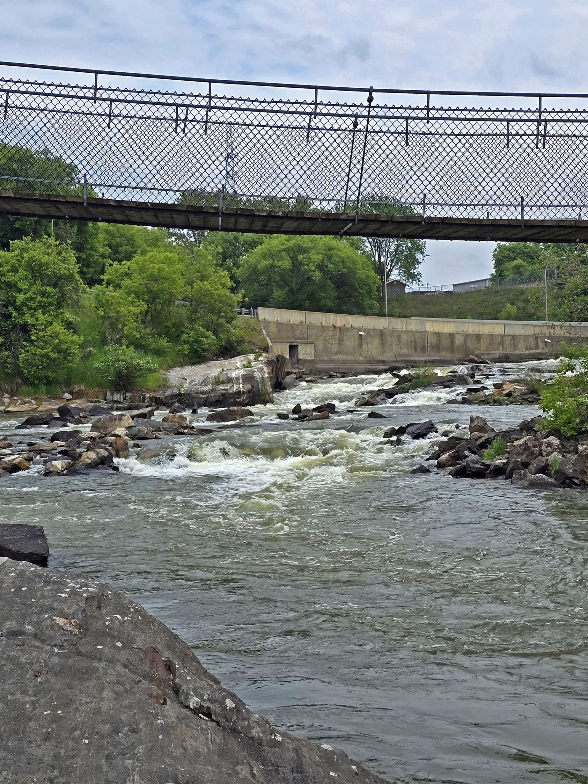 The Roy Wilson suspension bridge as seen from below and the side, with a small waterfall in the background.