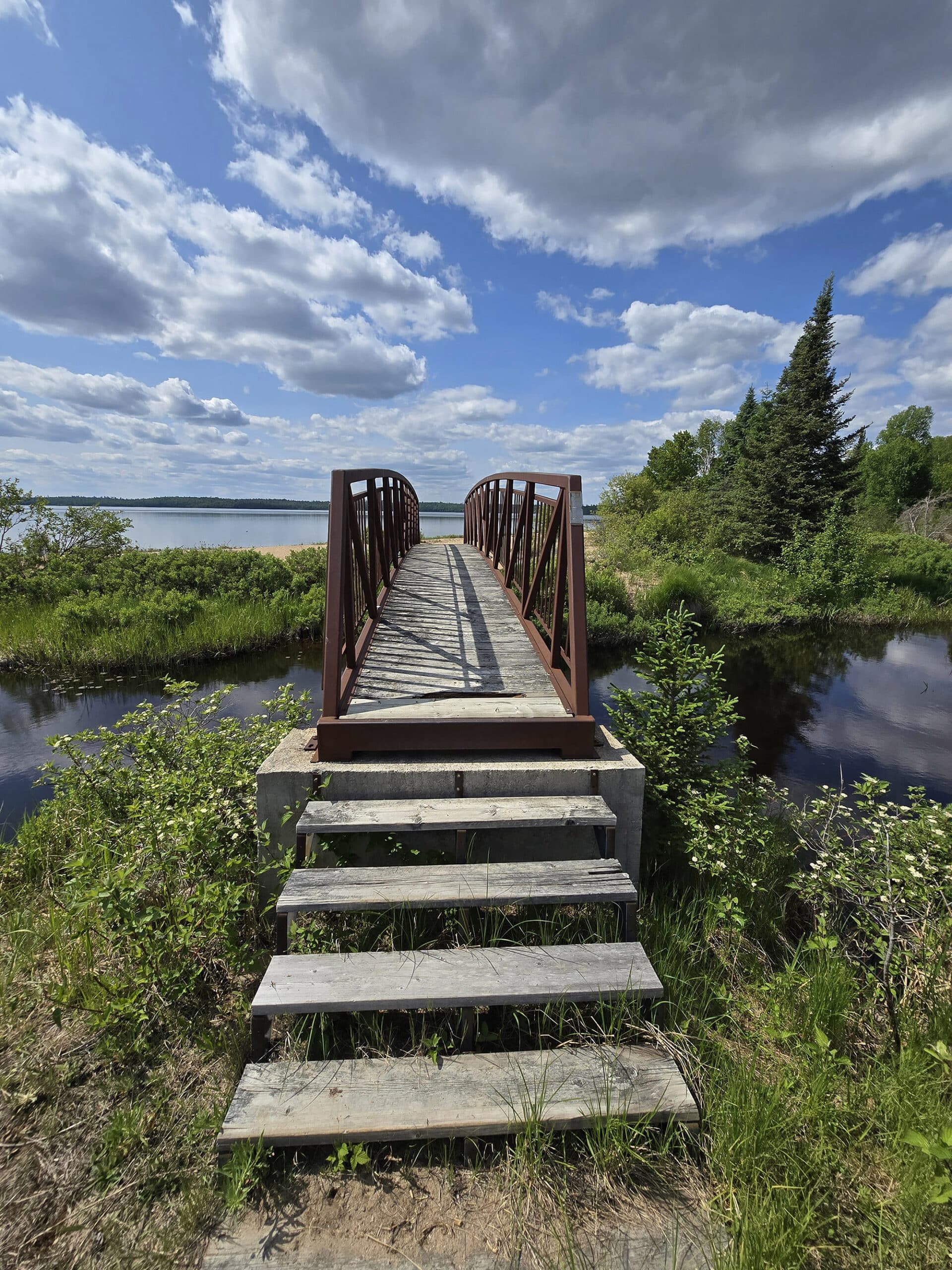 A footbridge over some water, going towards a sandy beach.