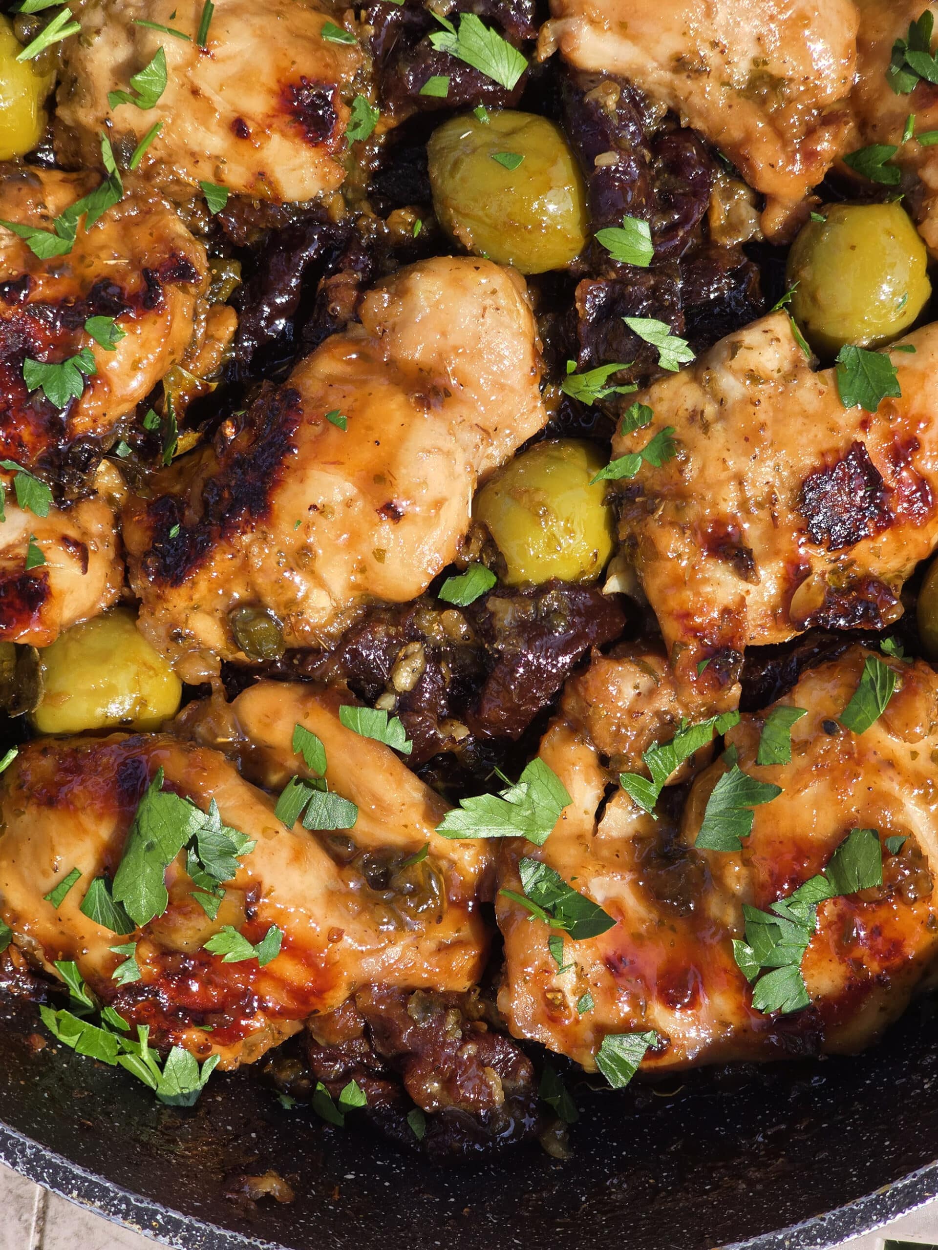 A pan of campfire chicken marbella, with caramelized chicken thighs, olives, prunes, and parsley visible.