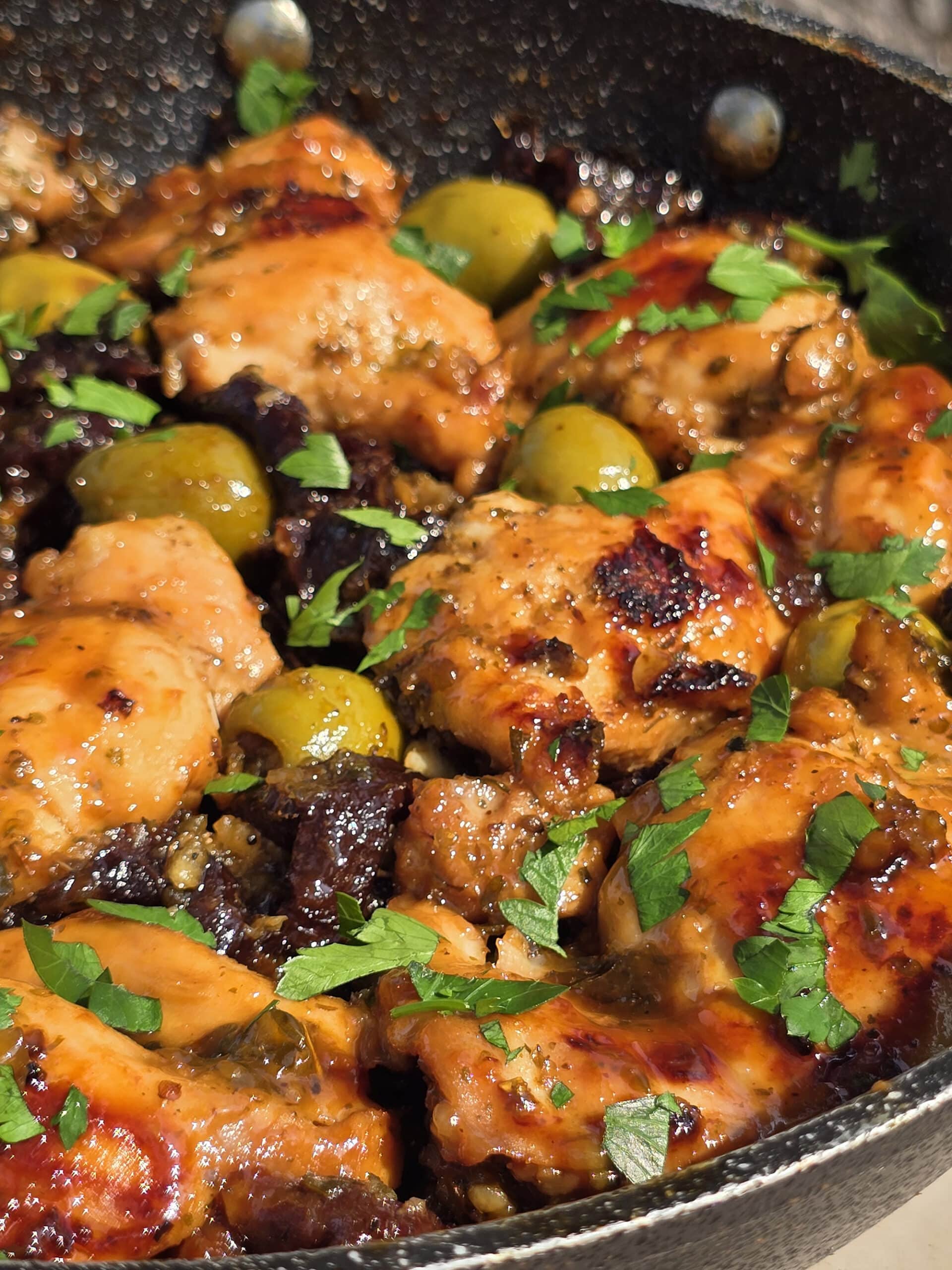 A pan of skillet chicken marbella, with caramelized chicken thighs, olives, prunes, and parsley visible.