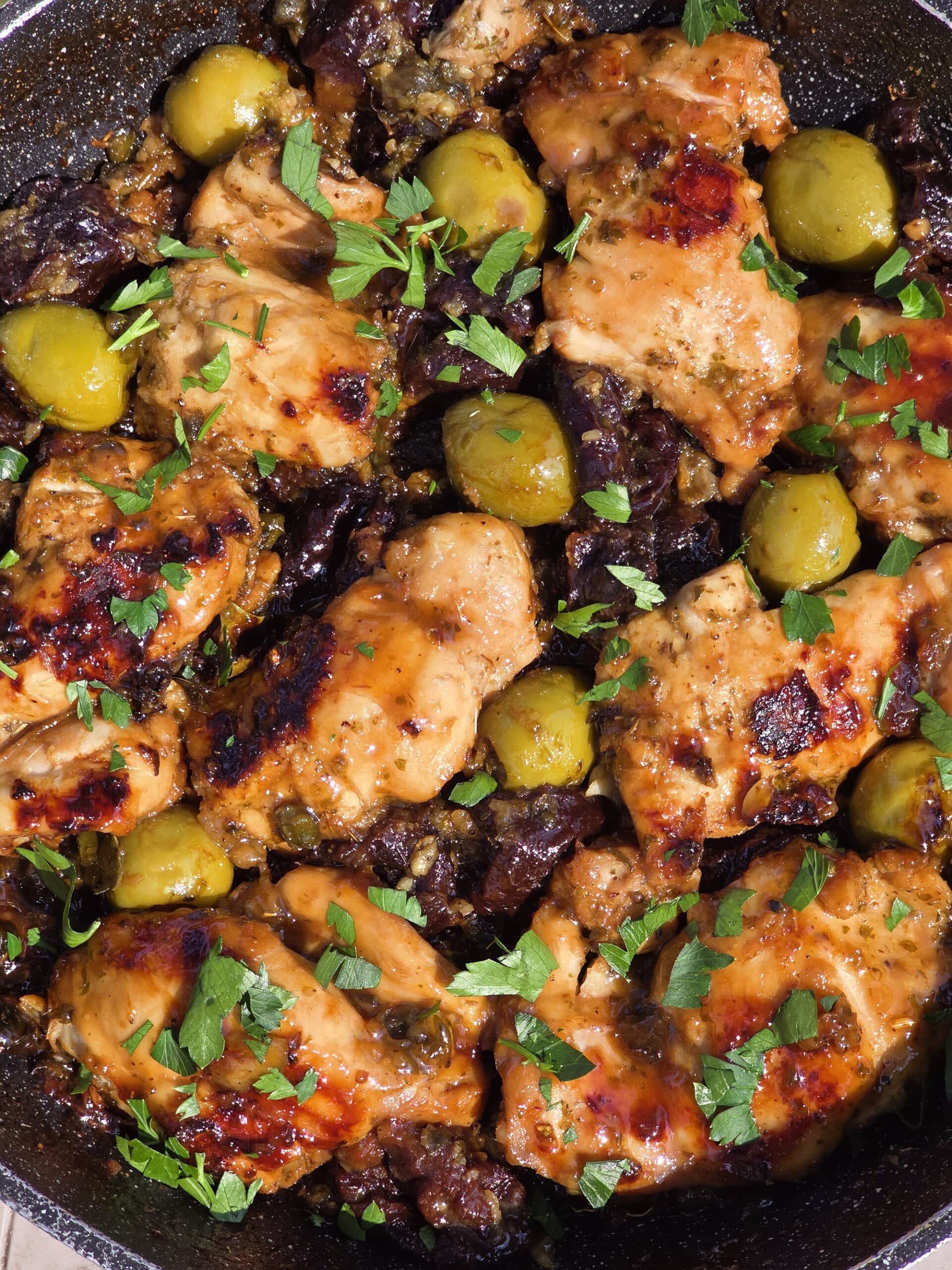 A pan of campfire chicken marbella, with caramelized chicken thighs, olives, prunes, and parsley visible.
