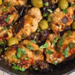 A pan of skillet chicken marbella, with caramelized chicken thighs, olives, prunes, and parsley visible.