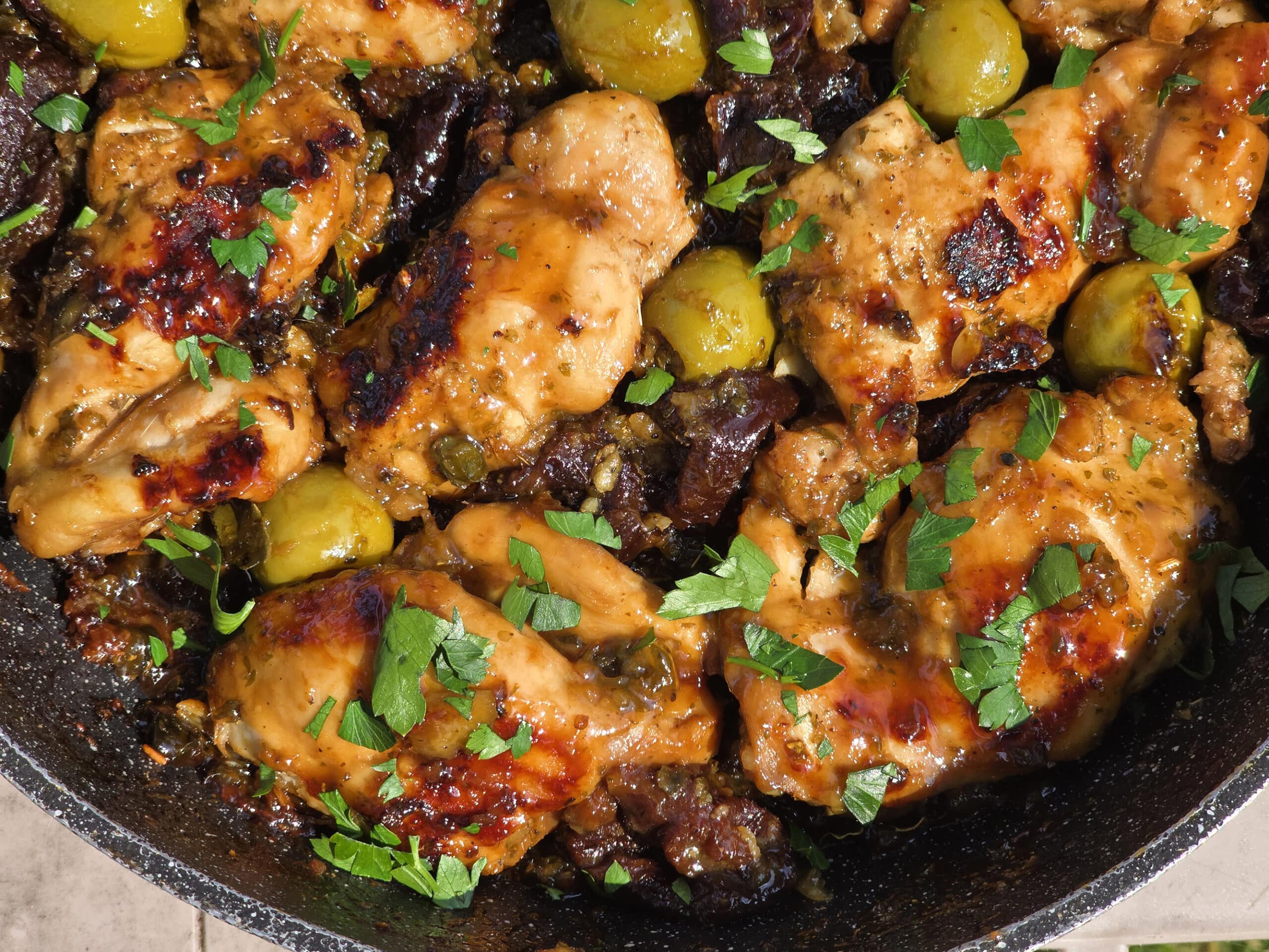 A pan of skillet chicken marbella, with caramelized chicken thighs, olives, prunes, and parsley visible.