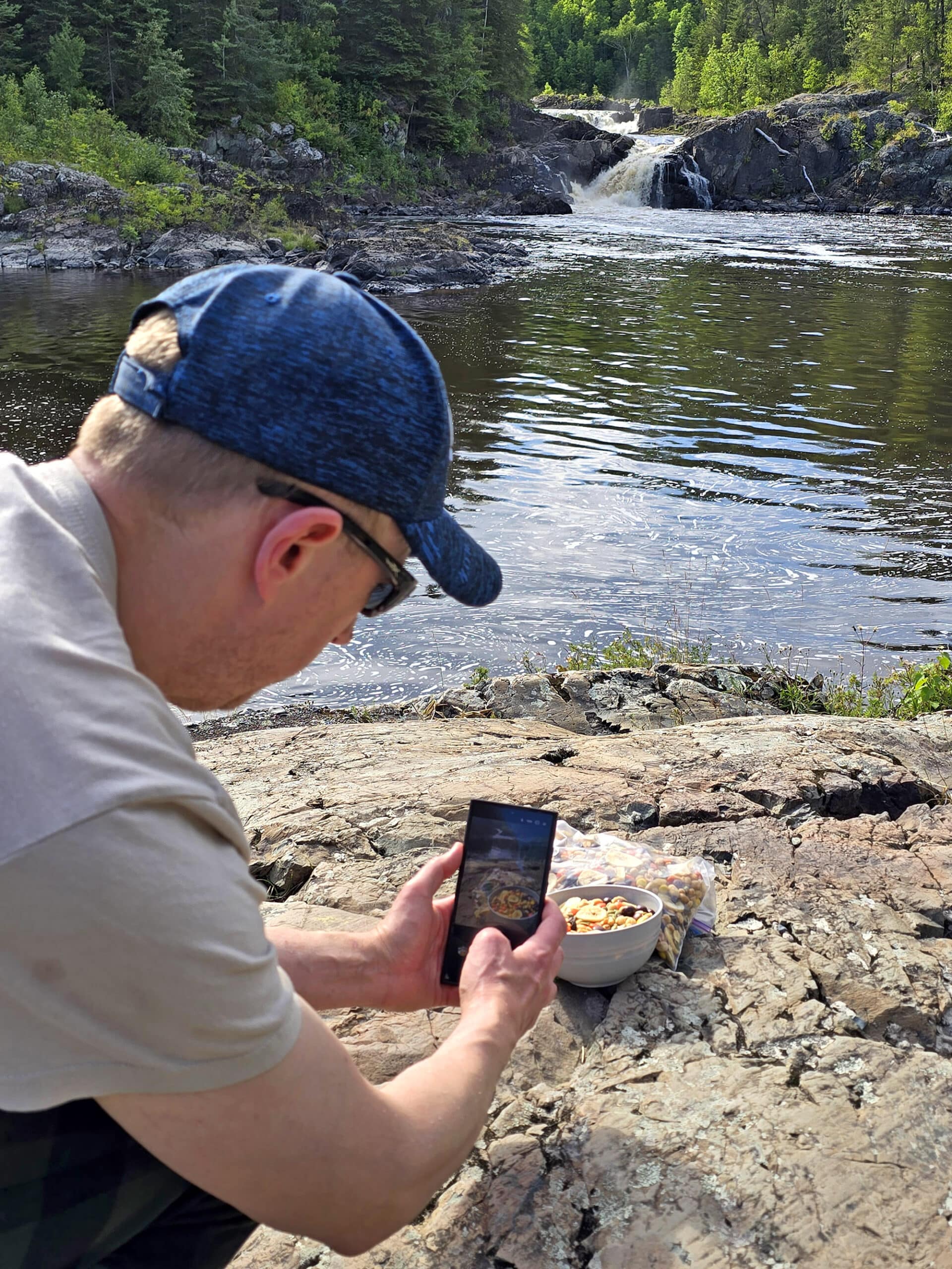 A man photographing a bowl of caffeinated trail mix on a rock in front of a waterfall.