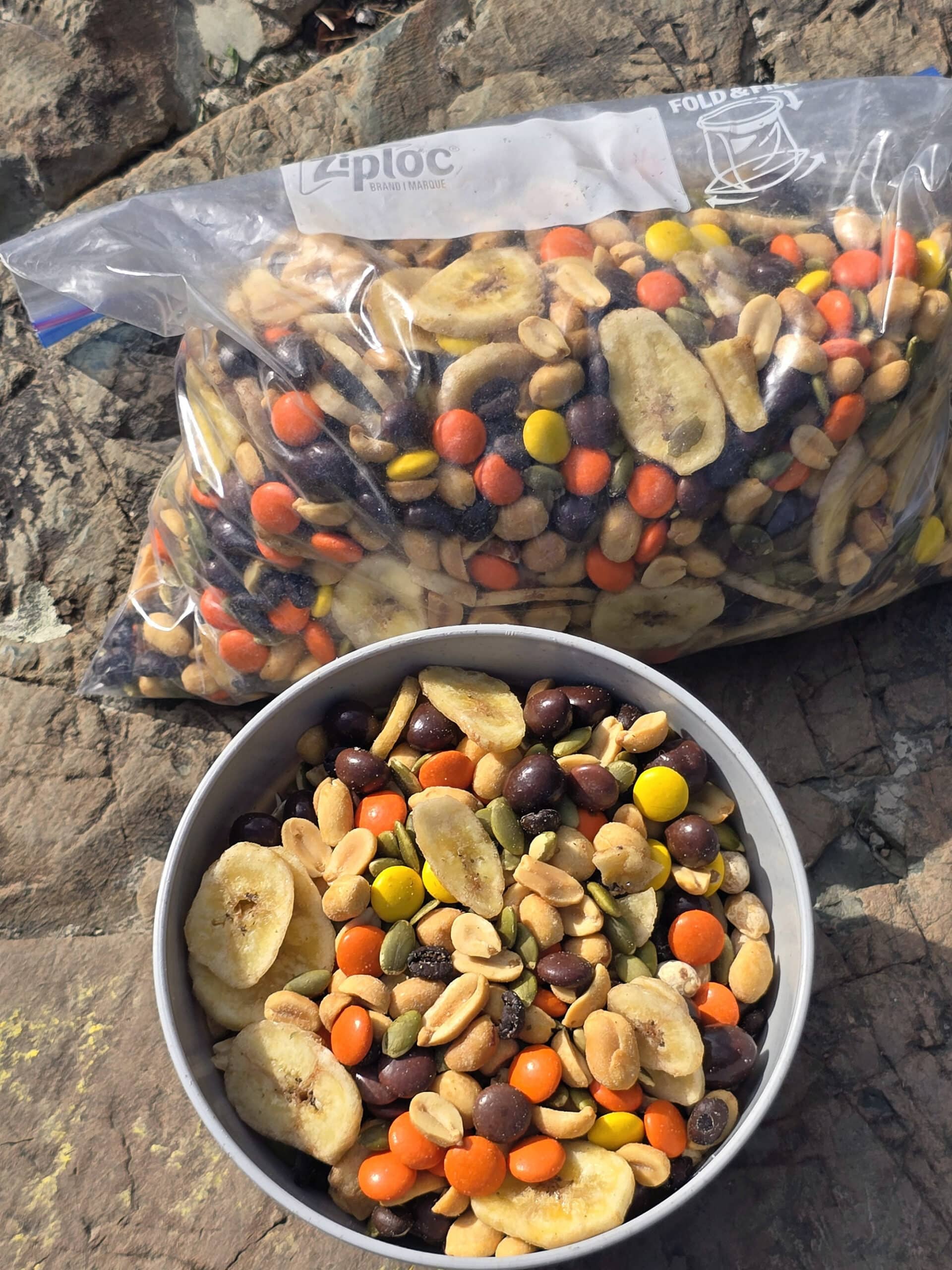 A bowl and bag of caffeinated trail mix with reeses pieces, chocolate covered coffee beans, pepitas, peanuts, and dried banana chips.