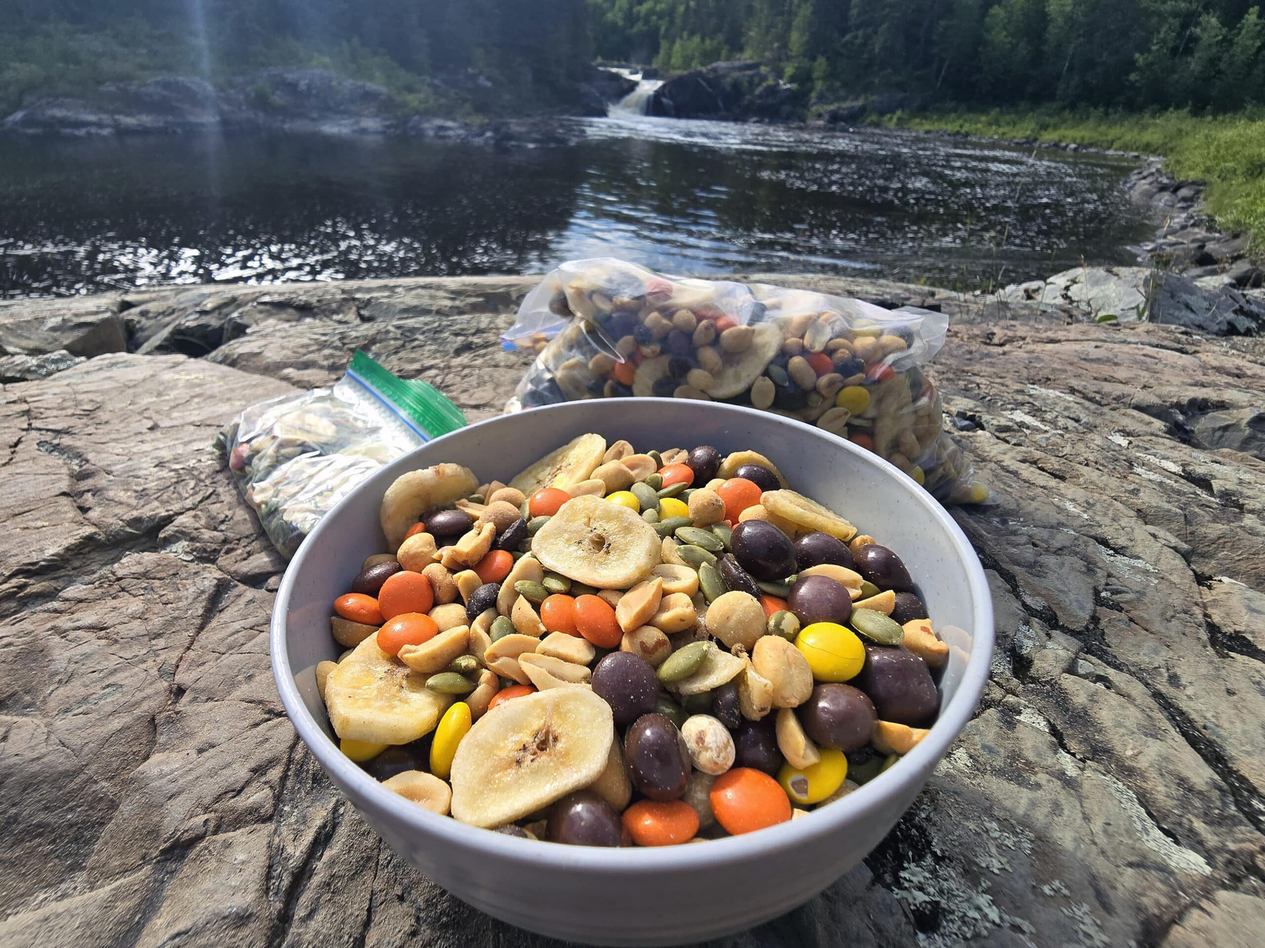 A bowl and bag of caffeinated trail mix with reeses pieces, chocolate covered coffee beans, pepitas, peanuts, and dried banana chips.
