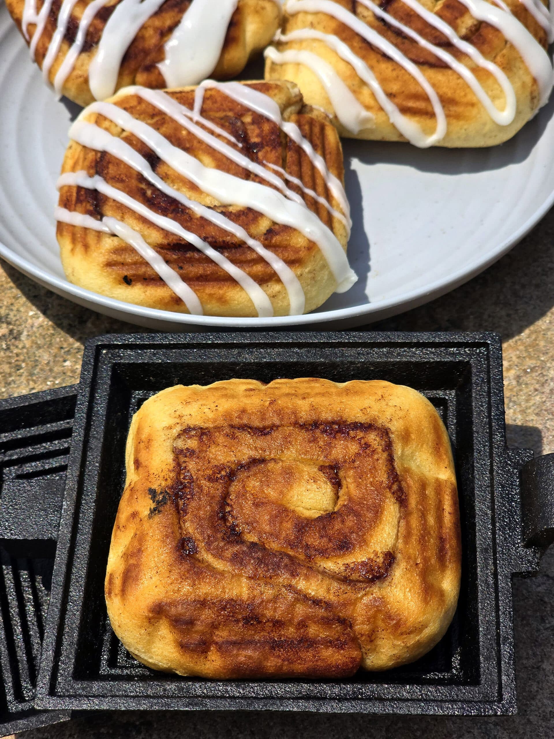 A plate of frosted pie iron cinnamon buns and an unfrosted cinnamon bun in a pie iron.