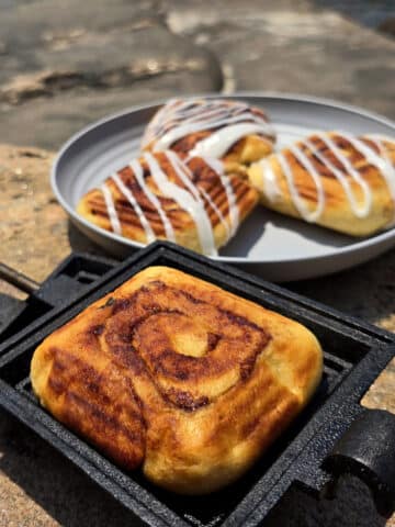 A plate of frosted pie iron cinnamon buns and an unfrosted cinnamon bun in a pie iron.