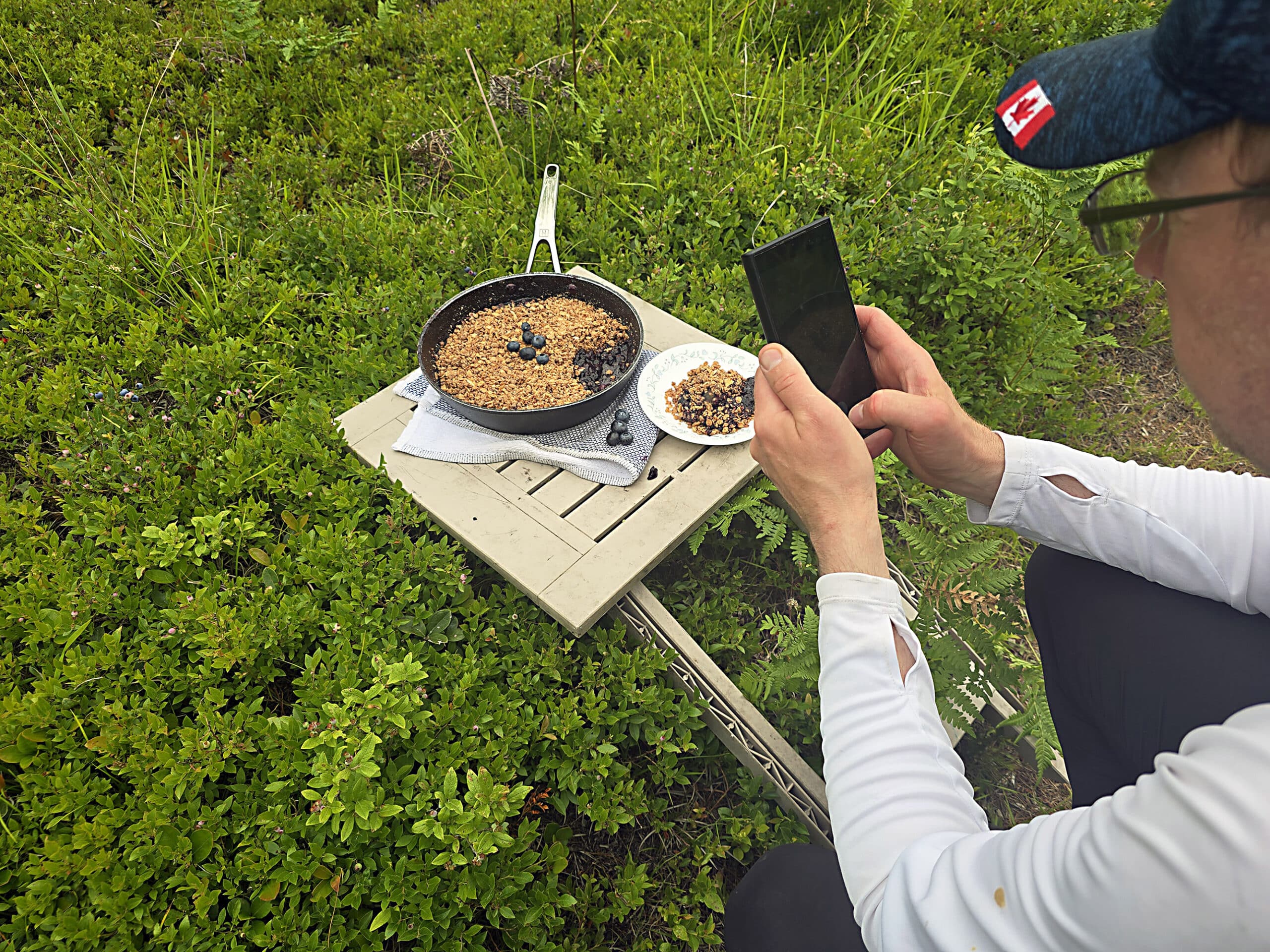 A man photographing a pan of blueberry crisp in a field of wild blueberry bushes.