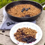 A pan of campfire blueberry crisp with a serving on a plate in front of it.
