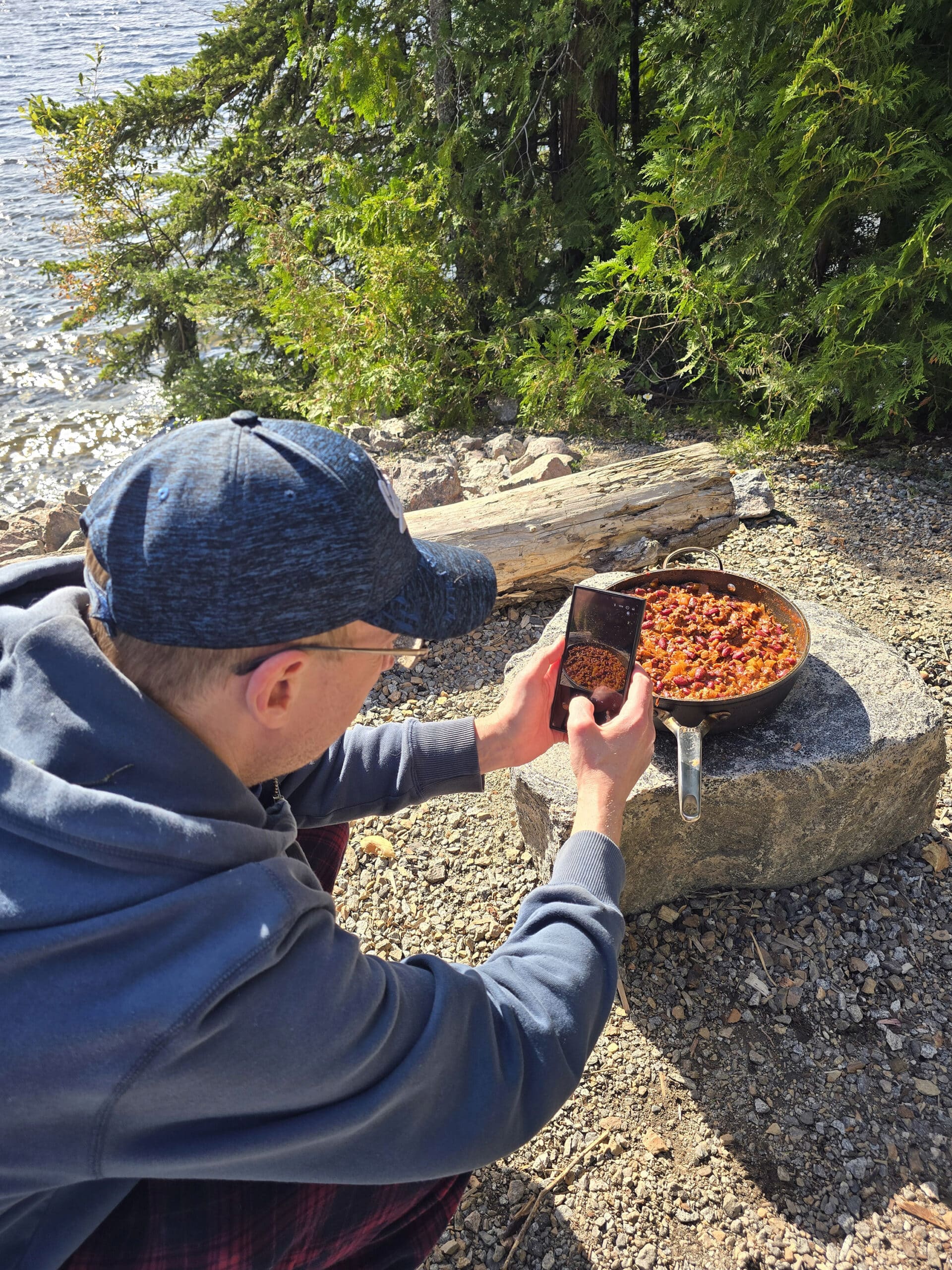 A pan in a hat photographing a pan of deer chili on a rock.