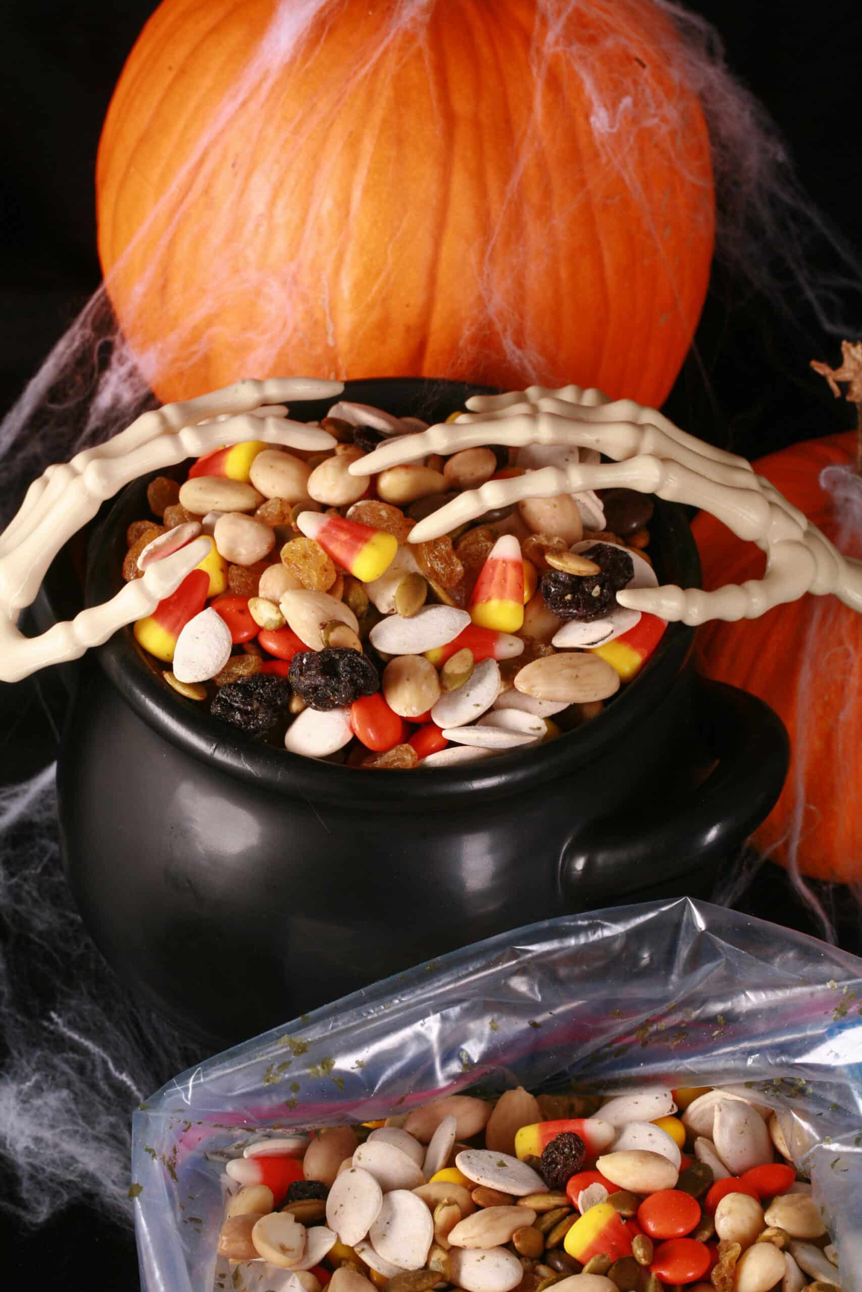 A small cauldron of halloween trail mix, with almonds, raisins, pumpkin seeds, candy corn, and more. There are pumpkins behind it, and skeleton hands posed over the cauldron.