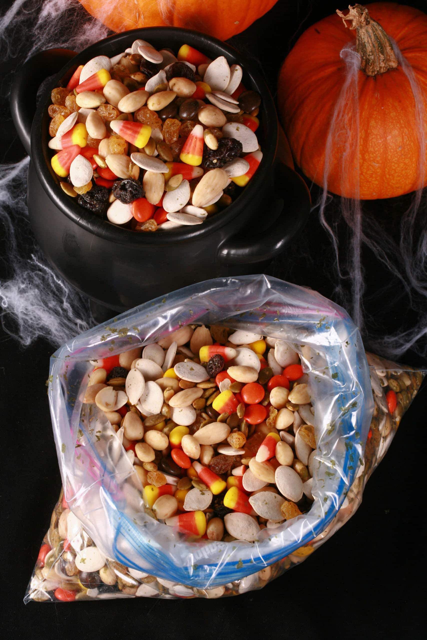 A small cauldron of halloween snack mix, with almonds, raisins, pumpkin seeds, candy corn, and more. There are pumpkins behind it, and a baggie of the same mix in front of it.