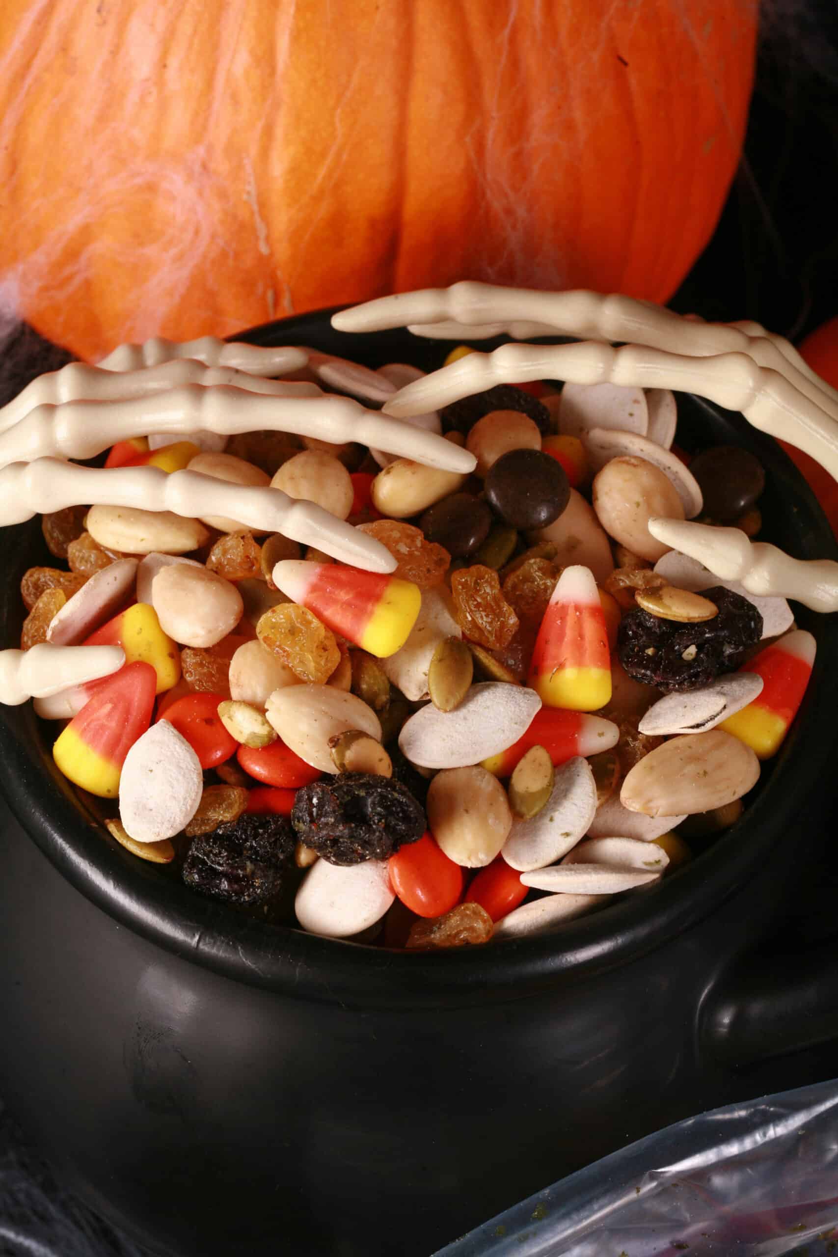 A small cauldron of halloween trail mix, with almonds, raisins, pumpkin seeds, candy corn, and more. There are pumpkins behind it, and skeleton hands posed over the cauldron.