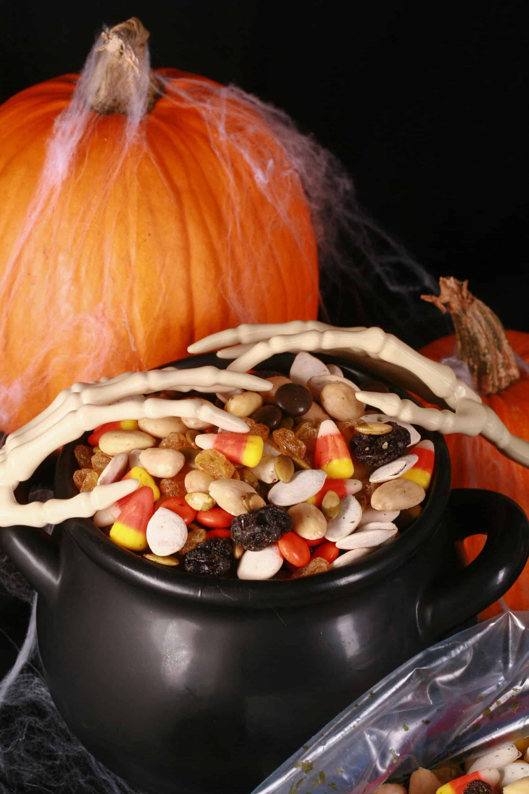 A small cauldron of halloween trail mix, with almonds, raisins, pumpkin seeds, candy corn, and more. There are pumpkins behind it, and skeleton hands posed over the cauldron.
