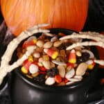 A small cauldron of halloween trail mix, with almonds, raisins, pumpkin seeds, candy corn, and more. There are pumpkins behind it, and skeleton hands posed over the cauldron.
