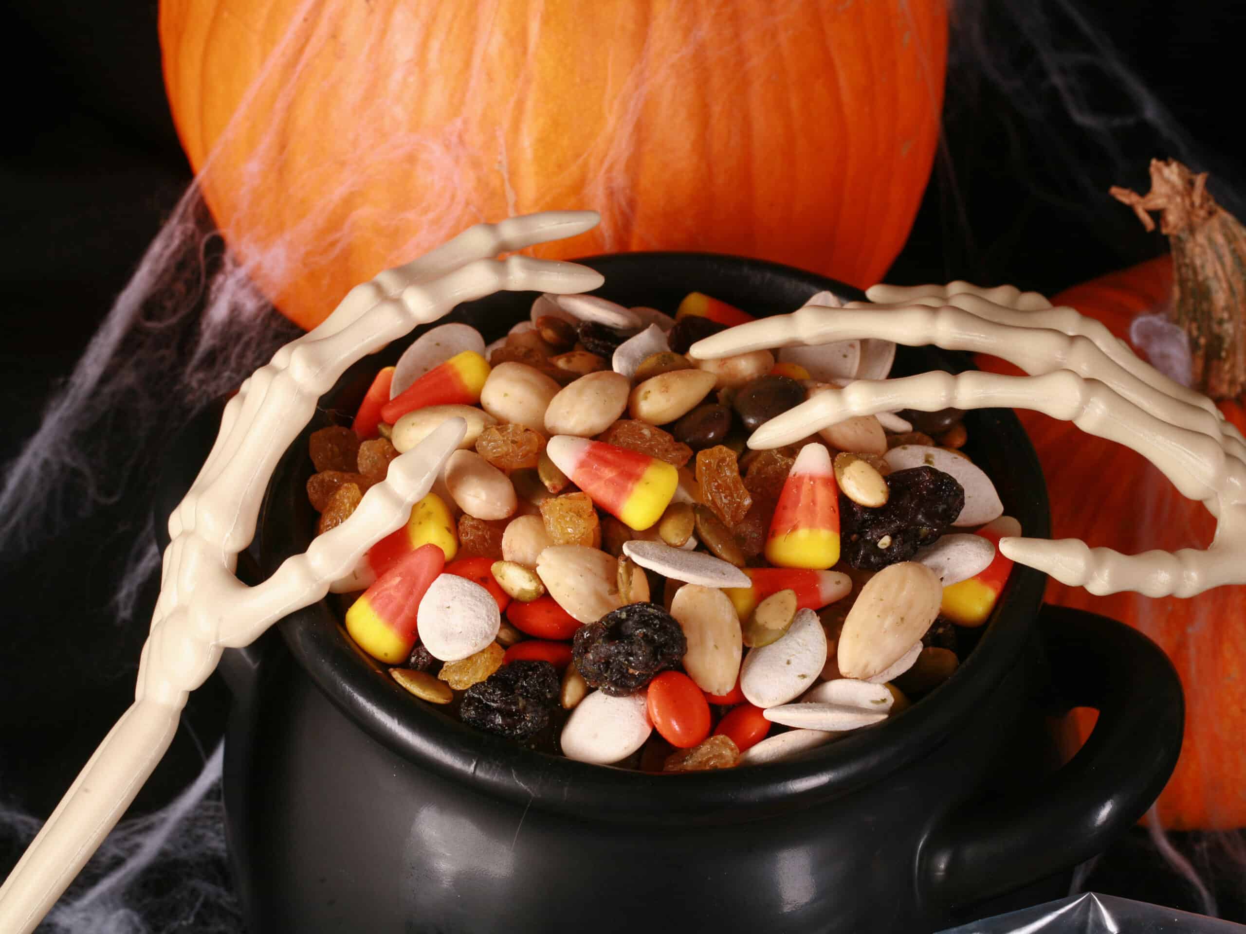A small cauldron of halloween trail mix, with almonds, raisins, pumpkin seeds, candy corn, and more. There are pumpkins behind it, and skeleton hands posed over the cauldron.