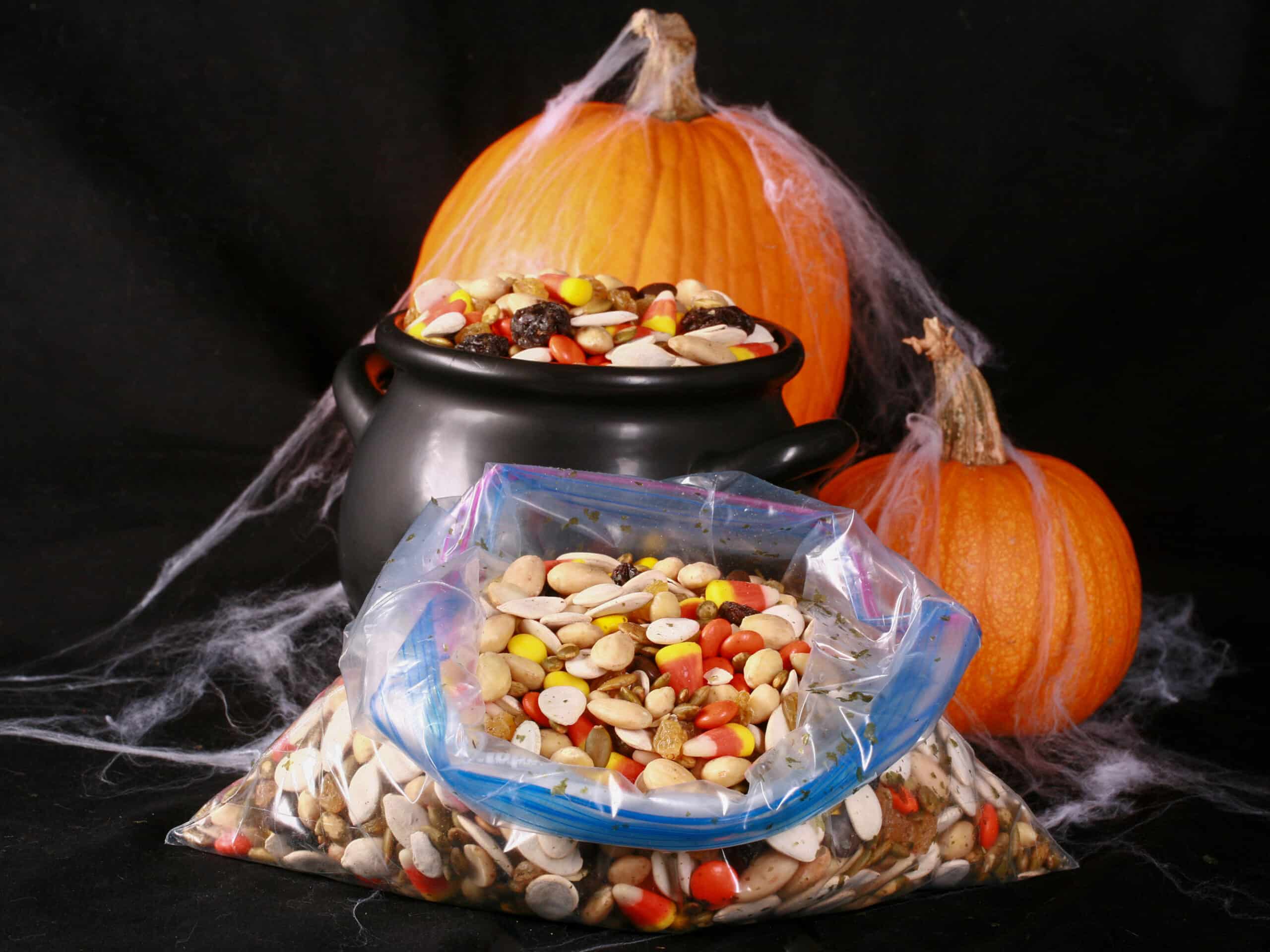 A small cauldron of halloween snack mix, with almonds, raisins, pumpkin seeds, candy corn, and more. There are pumpkins behind it, and a baggie of the same mix in front of it.