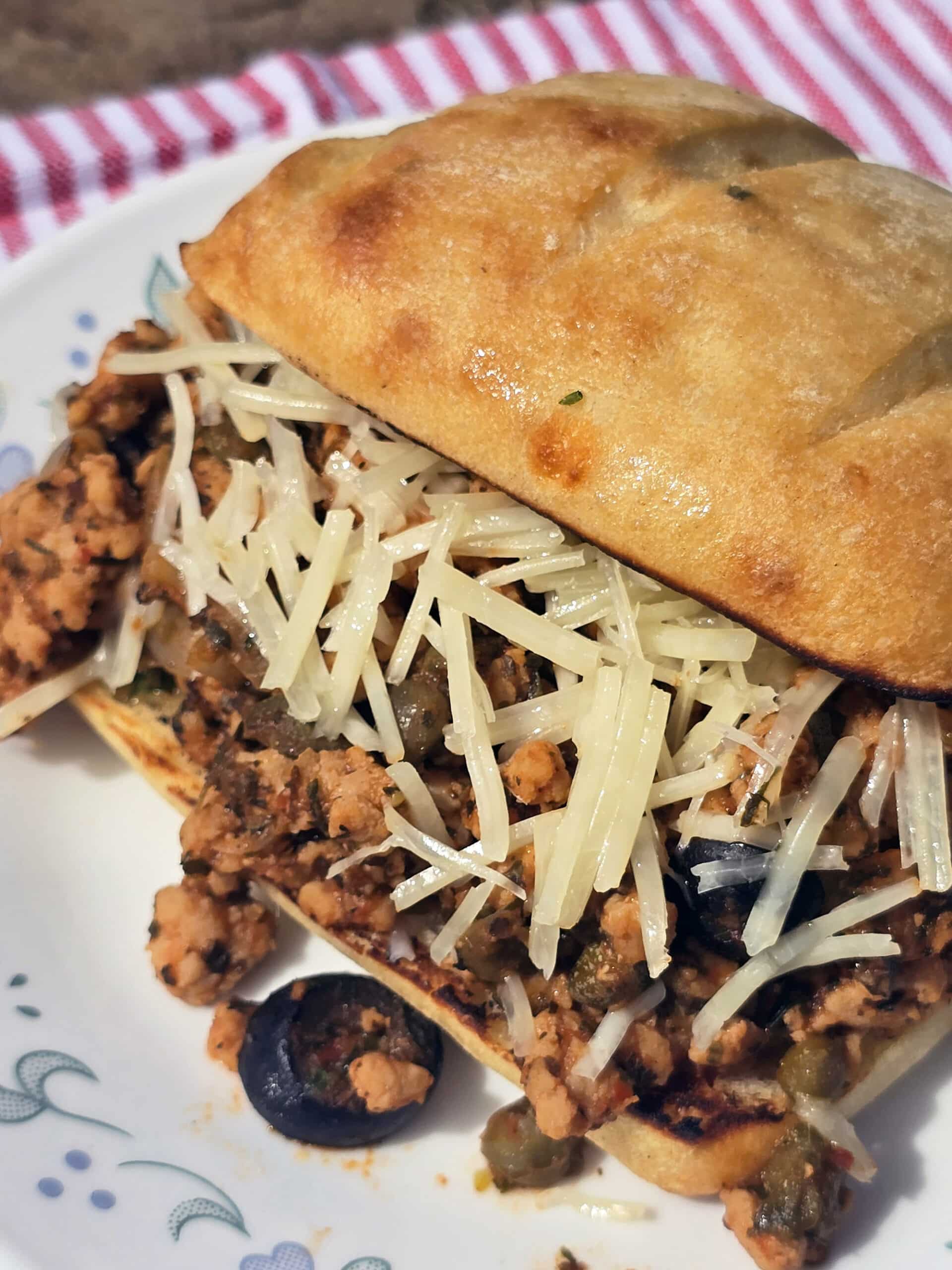 A Puttanesca sloppy joe on a ciabatta roll with black olives, capers, and parmesan cheese visible.