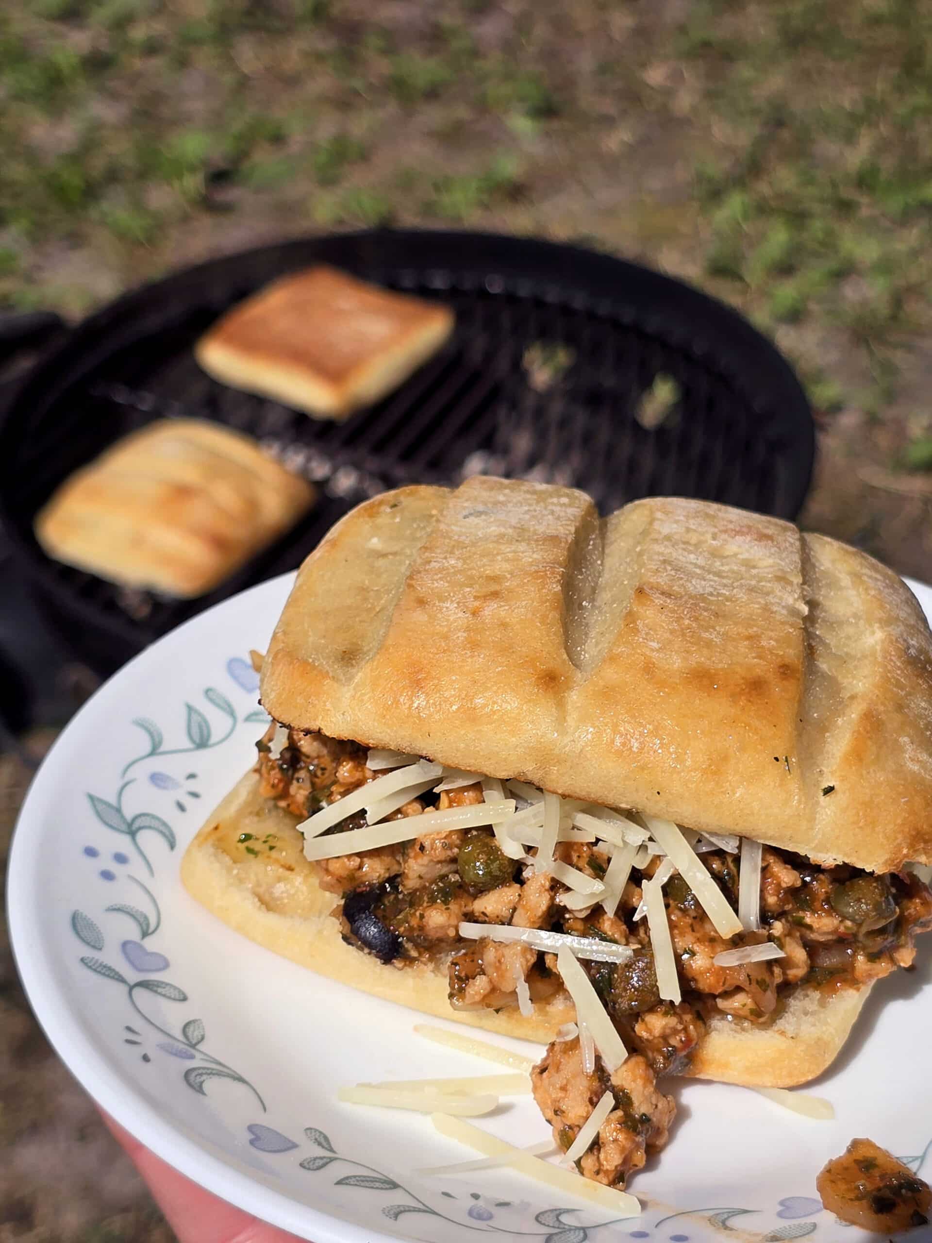 A Puttanesca sloppy joe on a ciabatta roll with black olives, capers, and parmesan cheese visible, and more ciabatta rolls grilling behind it.
