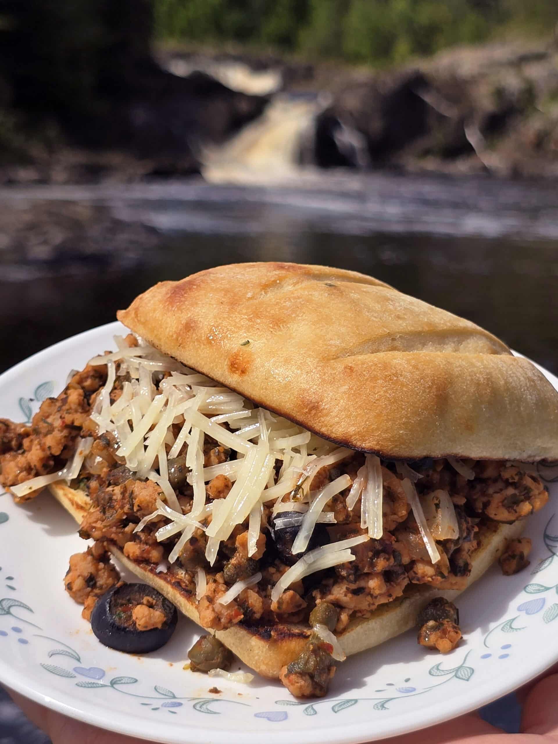 A Puttanesca sloppy joe on a ciabatta roll with black olives, capers, and parmesan cheese visible.