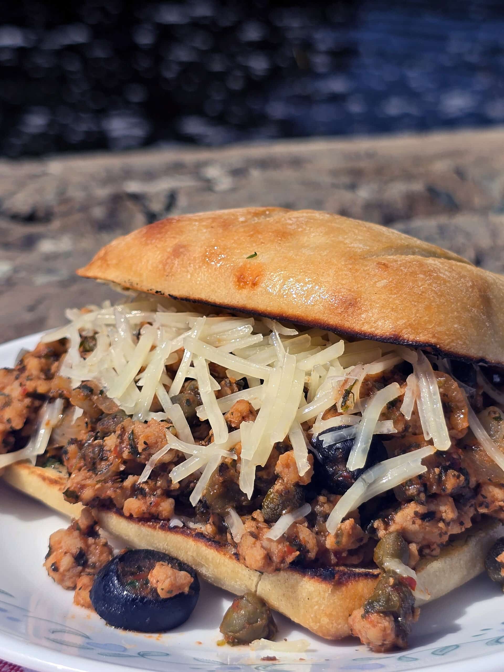 A Puttanesca sloppy joe on a ciabatta roll with black olives, capers, and parmesan cheese visible.