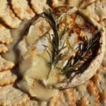 A round of smoky campfire brie with a sprig of rosemary and drizzle of maple syrup, surrounded by a ring of crackers.