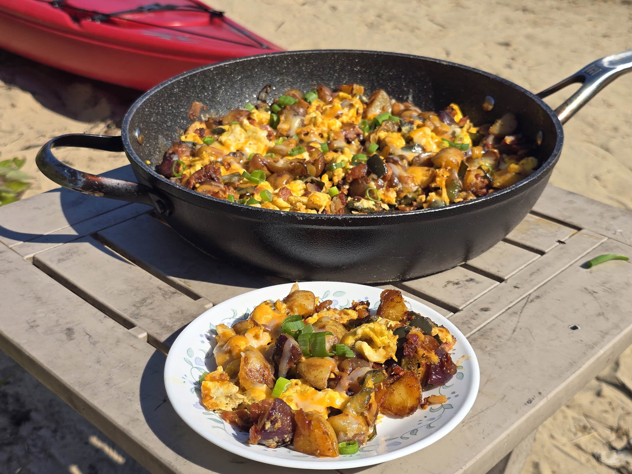 A pan and plate of smoky garlic chipotle breakfast skillet on a beach with kayaks behind it.