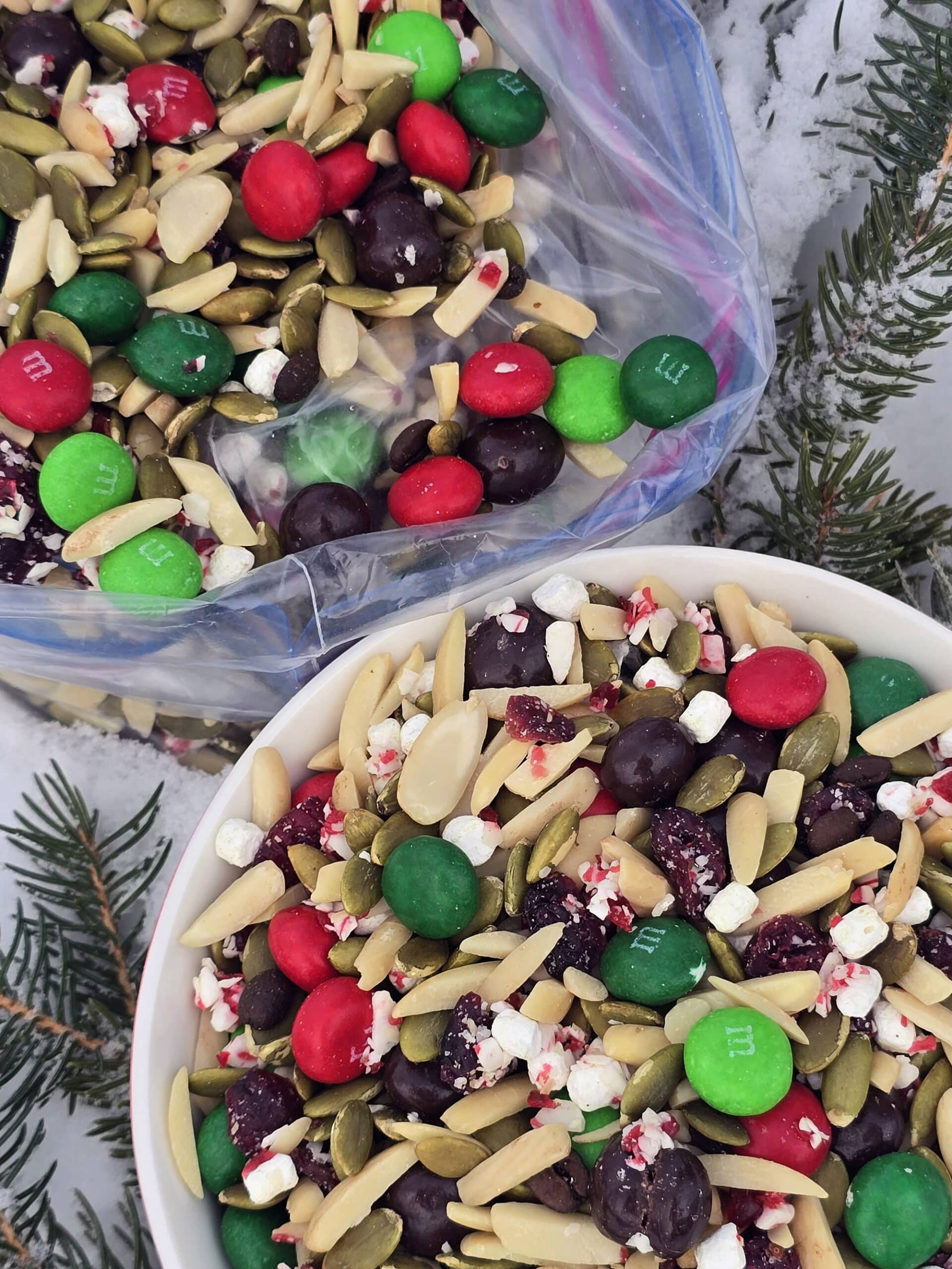 A bowl and a large plastic baggie of Christmas snack mix in the snow.