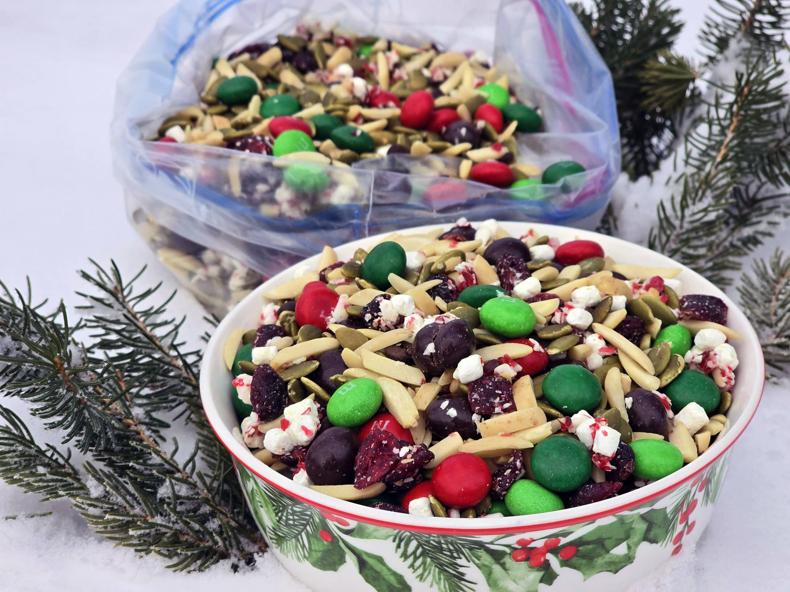 A bowl and a large plastic baggie of Christmas trail mix in the snow.