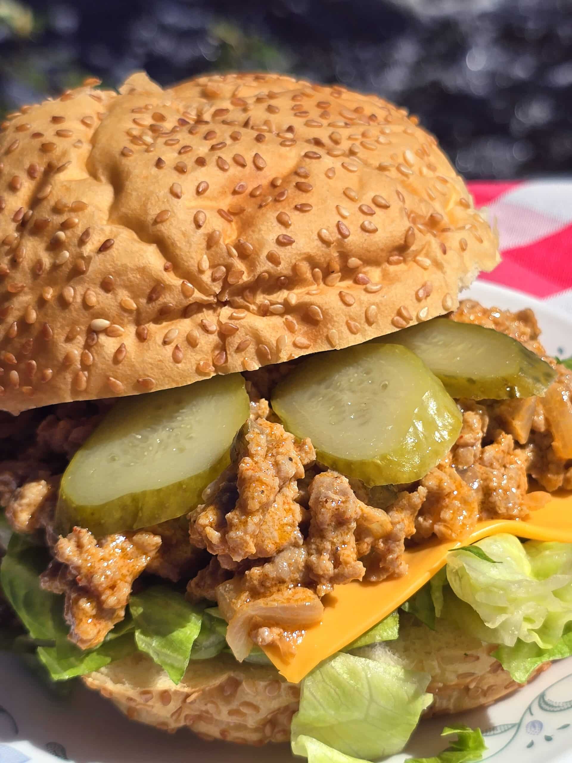A big mac sloppy joe sandwich on a plate in front of a waterfall. Lettuce, pickles, and cheese are visible.