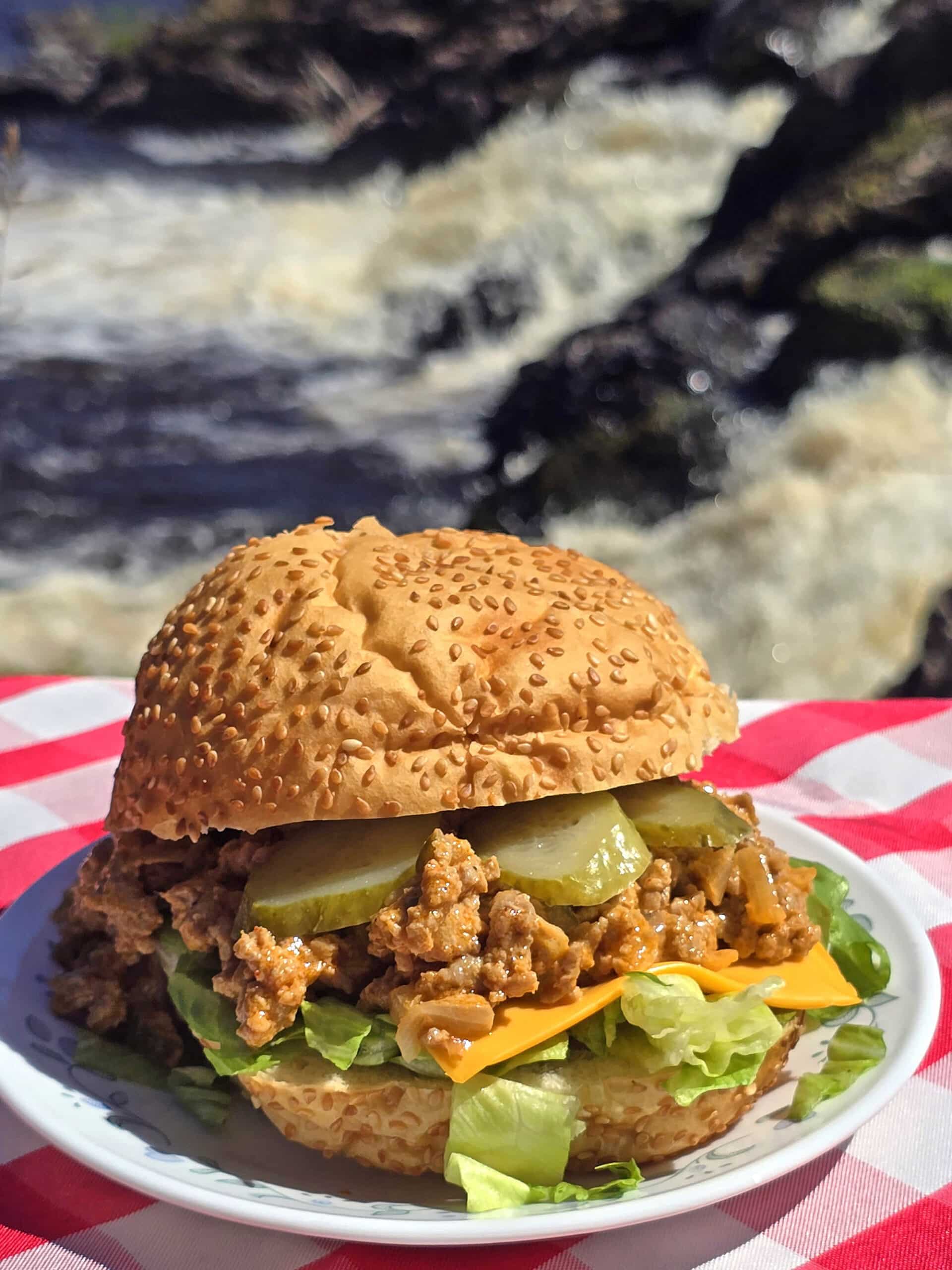 A big mac sloppy joe sandwich on a plate in front of a waterfall. Lettuce, pickles, and cheese are visible.