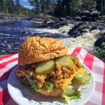 A big mac sloppy joe sandwich on a plate in front of a waterfall. Lettuce, pickles, and cheese are visible.