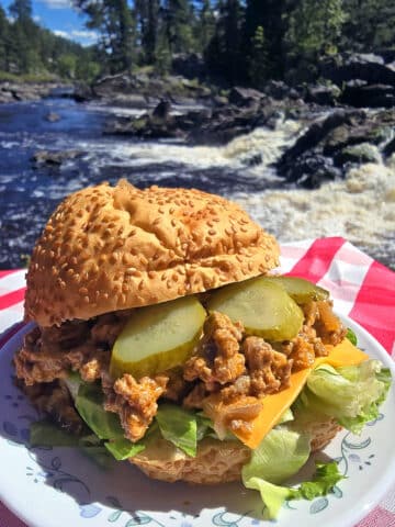 A big mac sloppy joe sandwich on a plate in front of a waterfall. Lettuce, pickles, and cheese are visible.