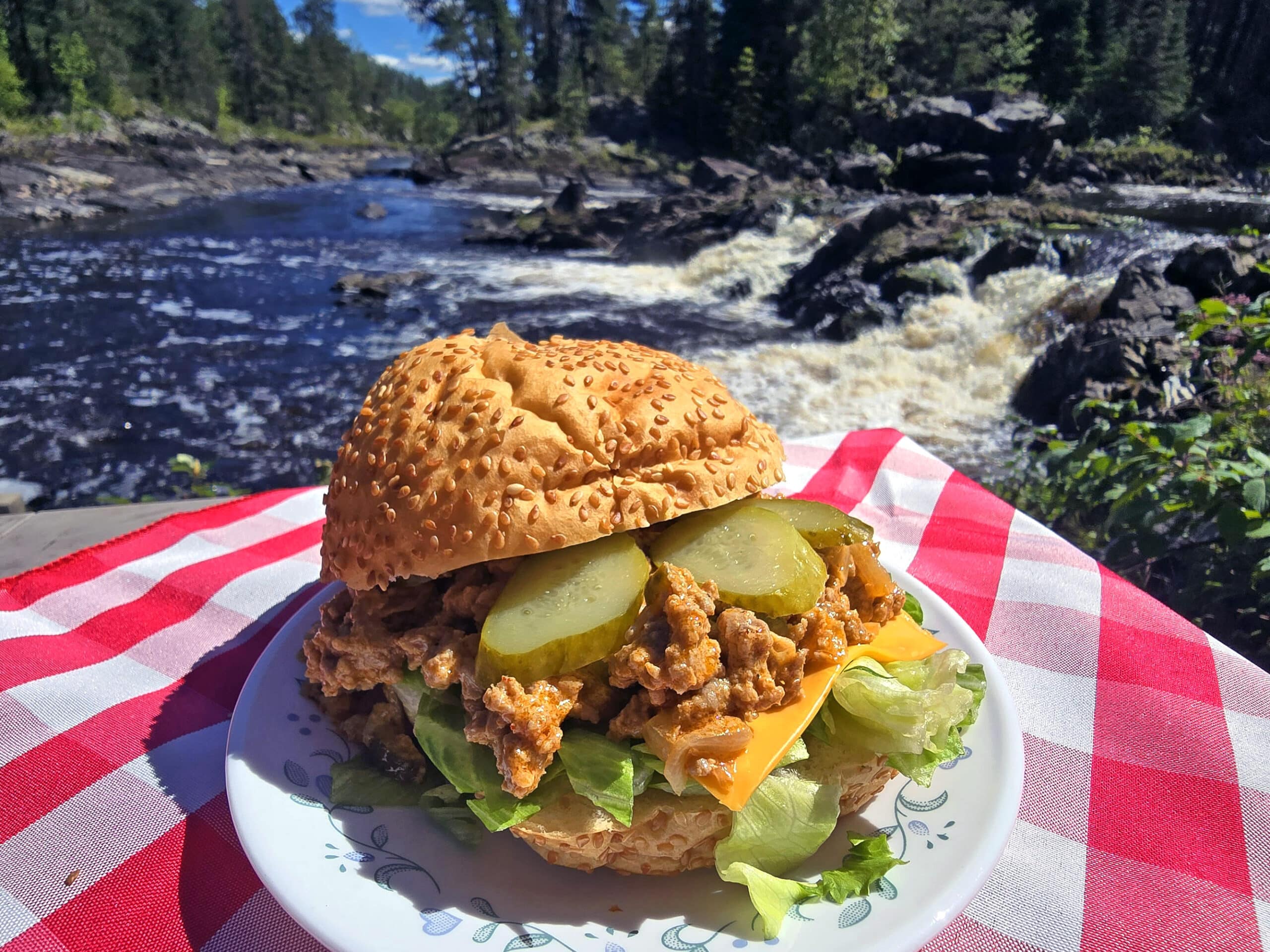 A big mac sloppy joe sandwich on a plate in front of a waterfall. Lettuce, pickles, and cheese are visible.