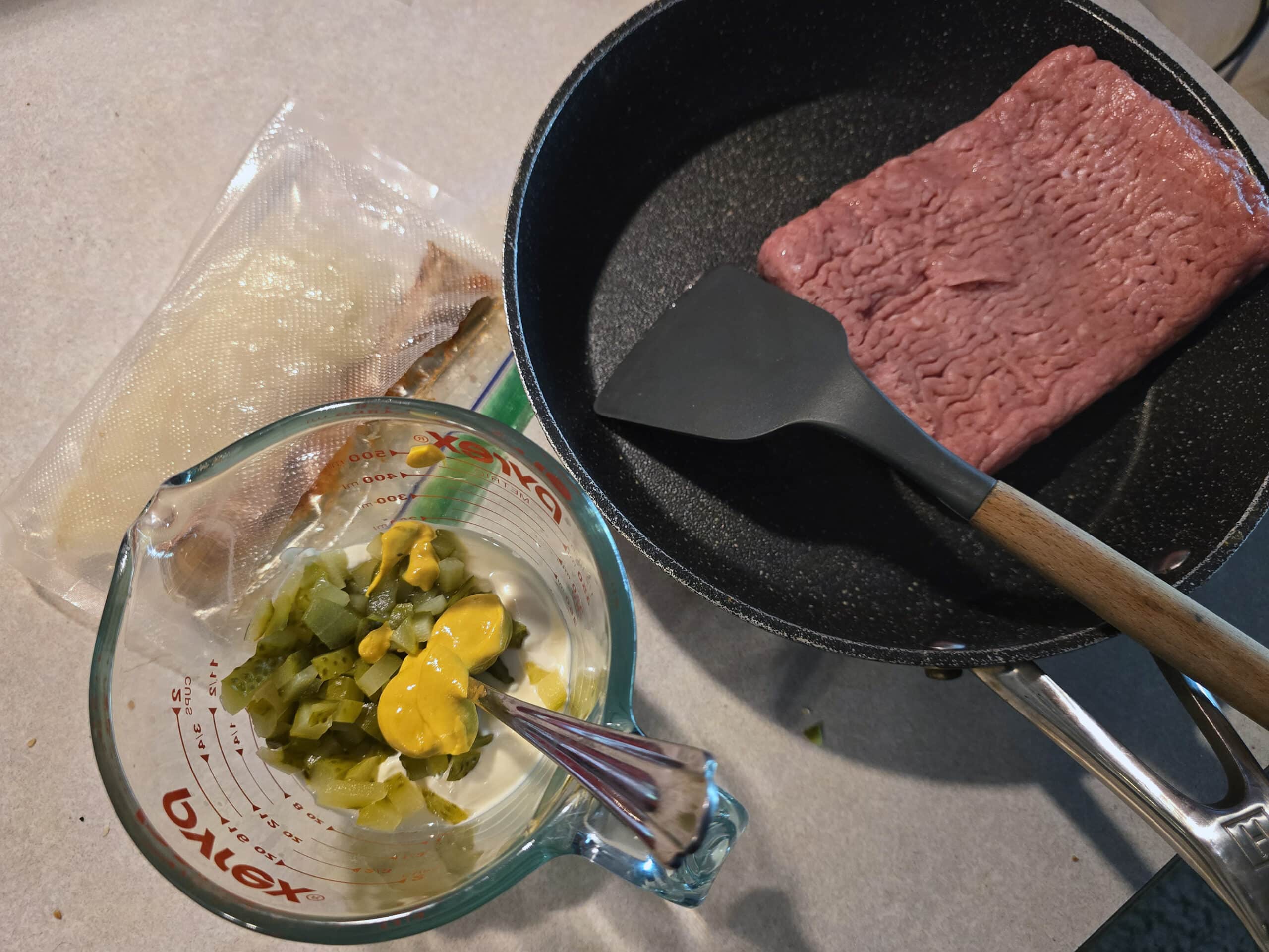 A pan of ground beef, a baggie with chopped onions and spices, and a glass bowl with mayo, mustard, and pickles in it.