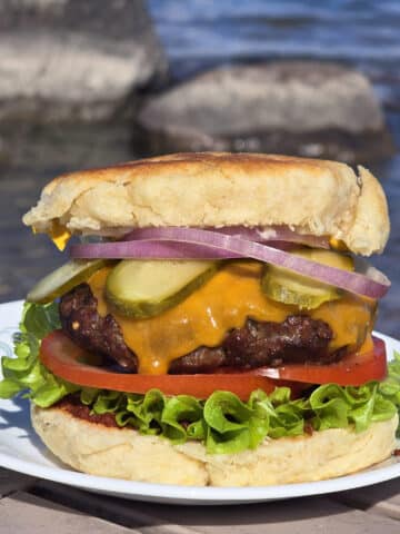 A bannock cheeseburger with bannock bun, beef patty, lettuce, tomato slices, cheddar cheese, pickle slices, and red onions.