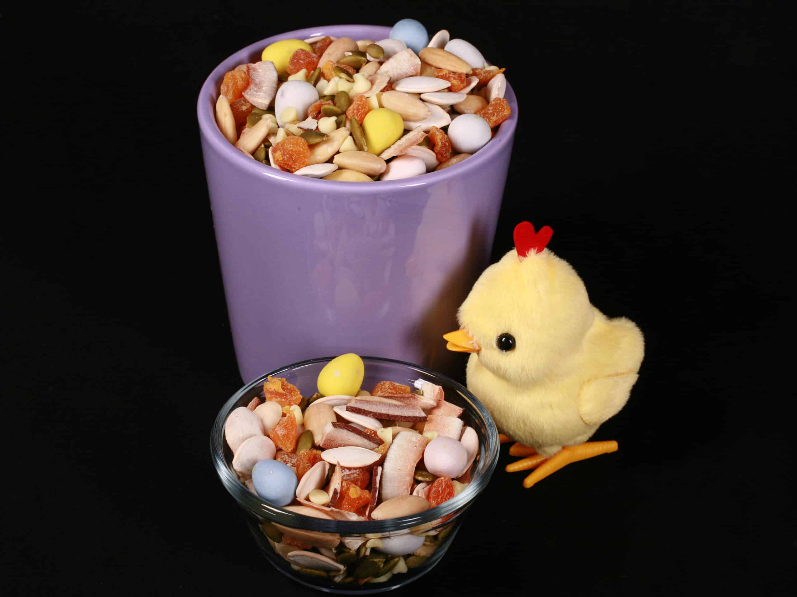 2 bowls of Easter trail mix, with coconut, apricot, pumpkin seeds, pepitas, which chocolate, and mini eggs. There is a toy chick next to them.