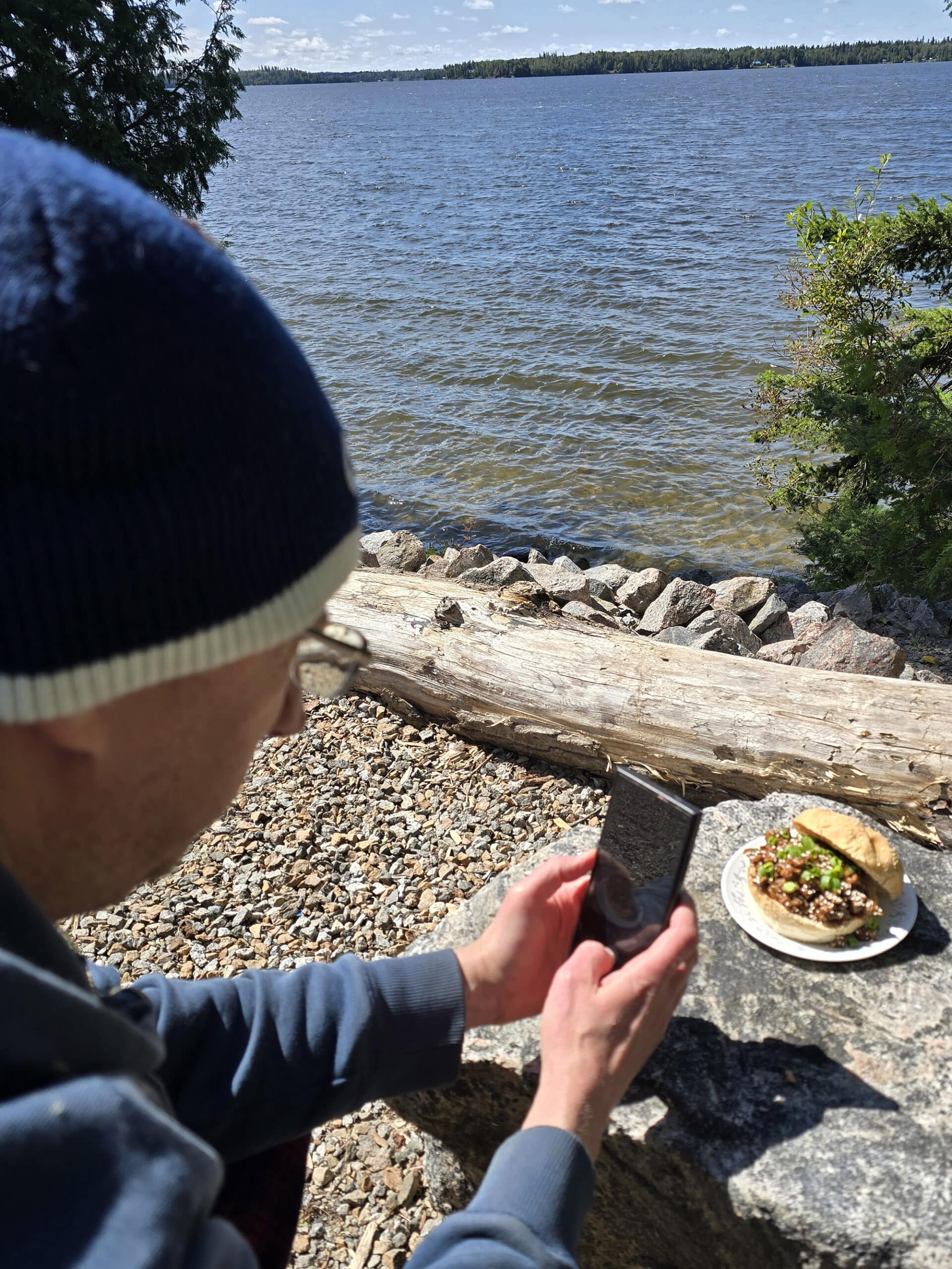 A man photographing a Korean BBQ sloppy joes sandwich next to a lake.