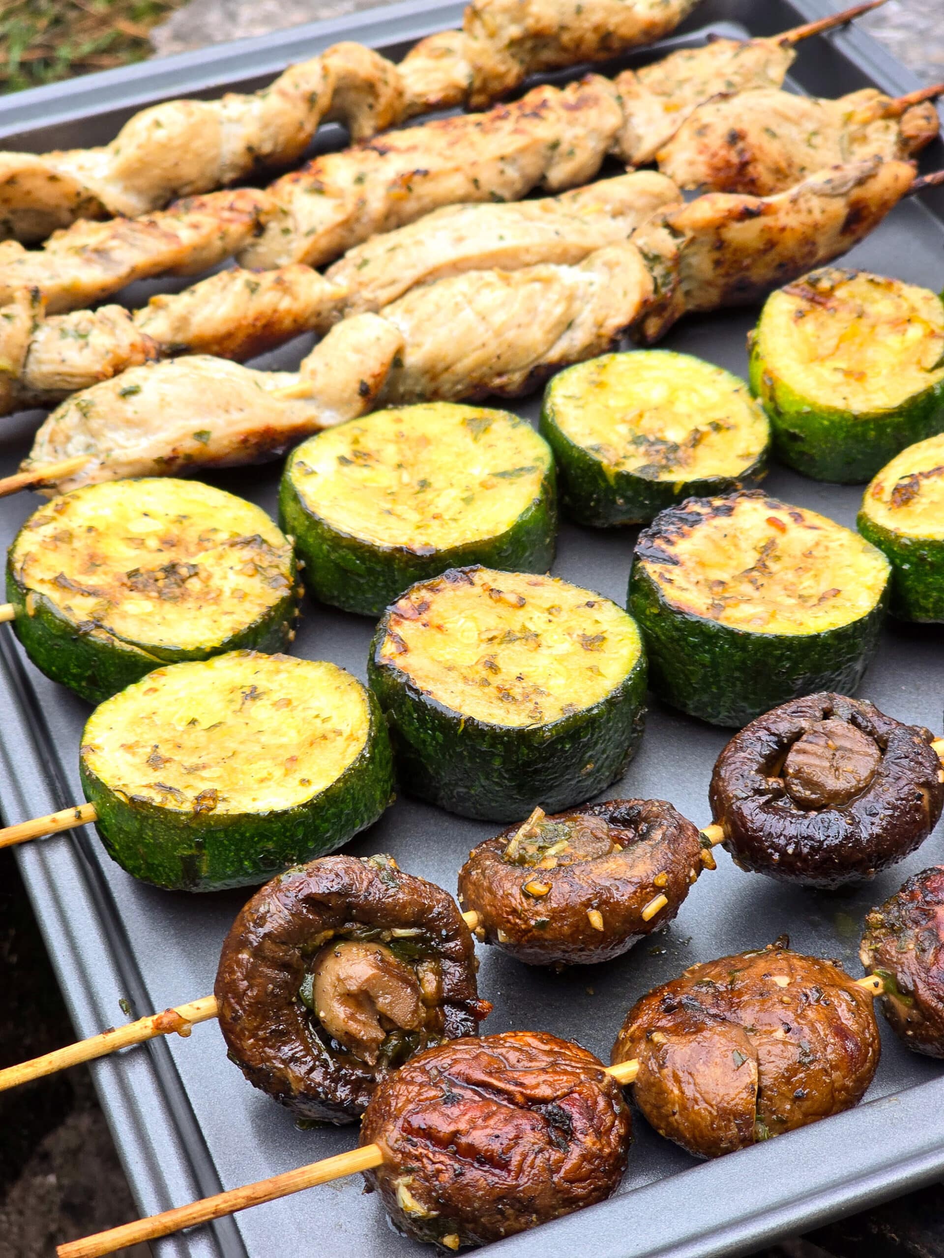 Individual skewers of pesto mushrooms and zucchini slices on a pan with pesto chicken kebabs.
