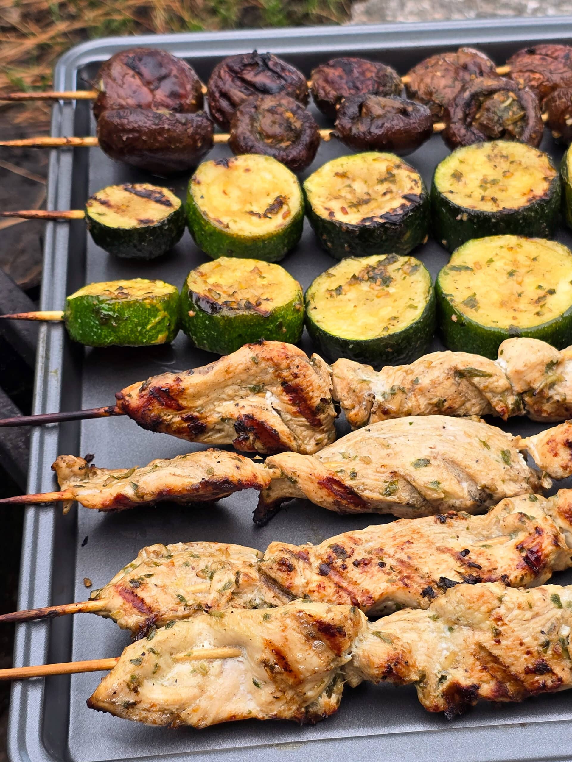 Individual skewers of pesto mushrooms and zucchini slices on a pan with pesto chicken kebabs.