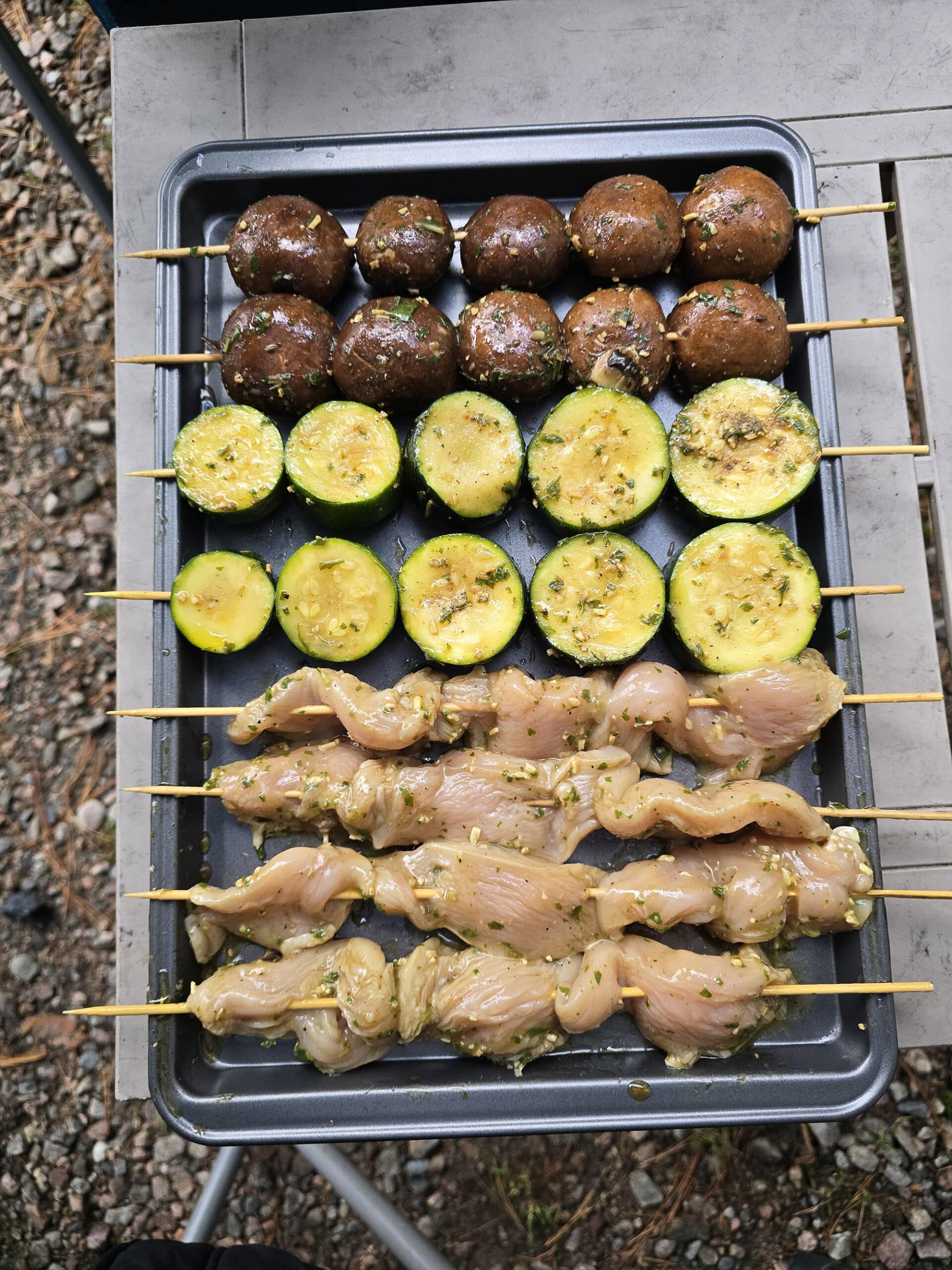 Individual skewers of marinated mushrooms, zucchini slices, and chicken laid out on a pan.