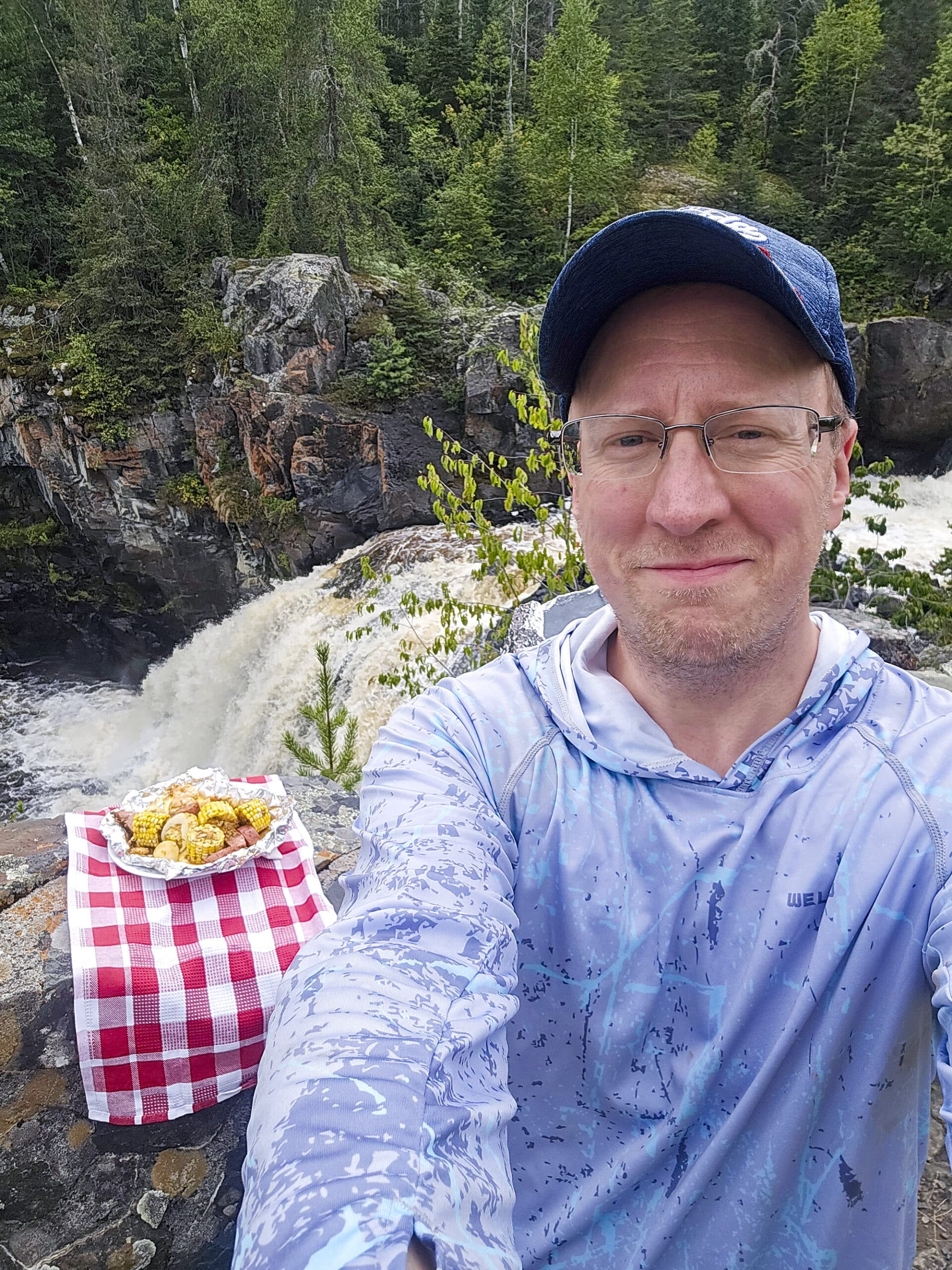 A man taking a selfie in front of a plate of shrimp boil foil packet and a waterfall.