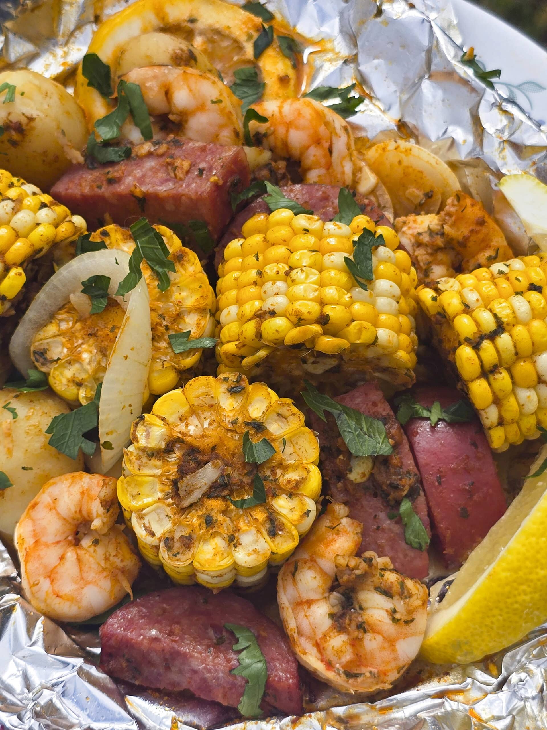 A large serving of seafood boil foil packet meal, on a plate. It has chunks of corn, sausage, potatoes, and shrimp in an obviously heavily seasoned sauce.