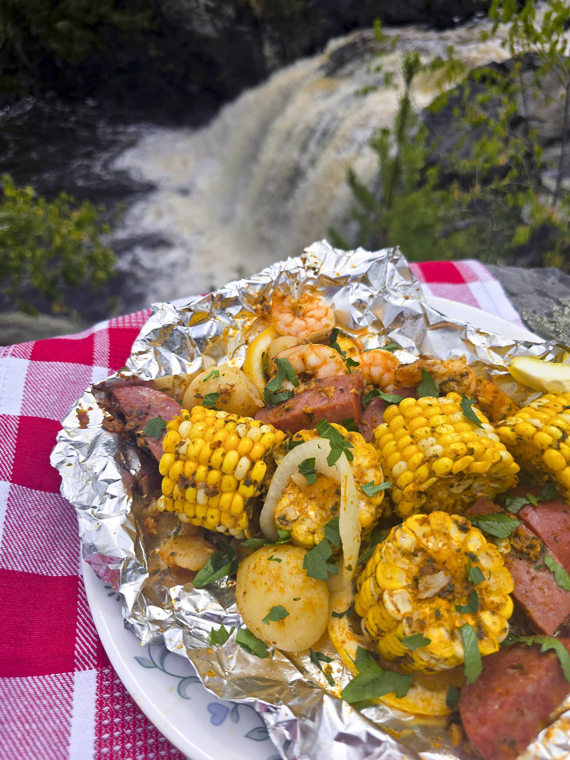 A large serving of shrimp boil foil packet meal, on a plate. It has chunks of corn, sausage, potatoes, and shrimp in an obviously heavily seasoned sauce.