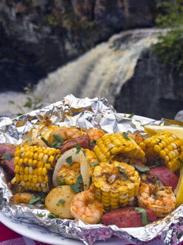 A large serving of seafood boil foil packet meal, on a plate. It has chunks of corn, sausage, potatoes, and shrimp in an obviously heavily seasoned sauce.
