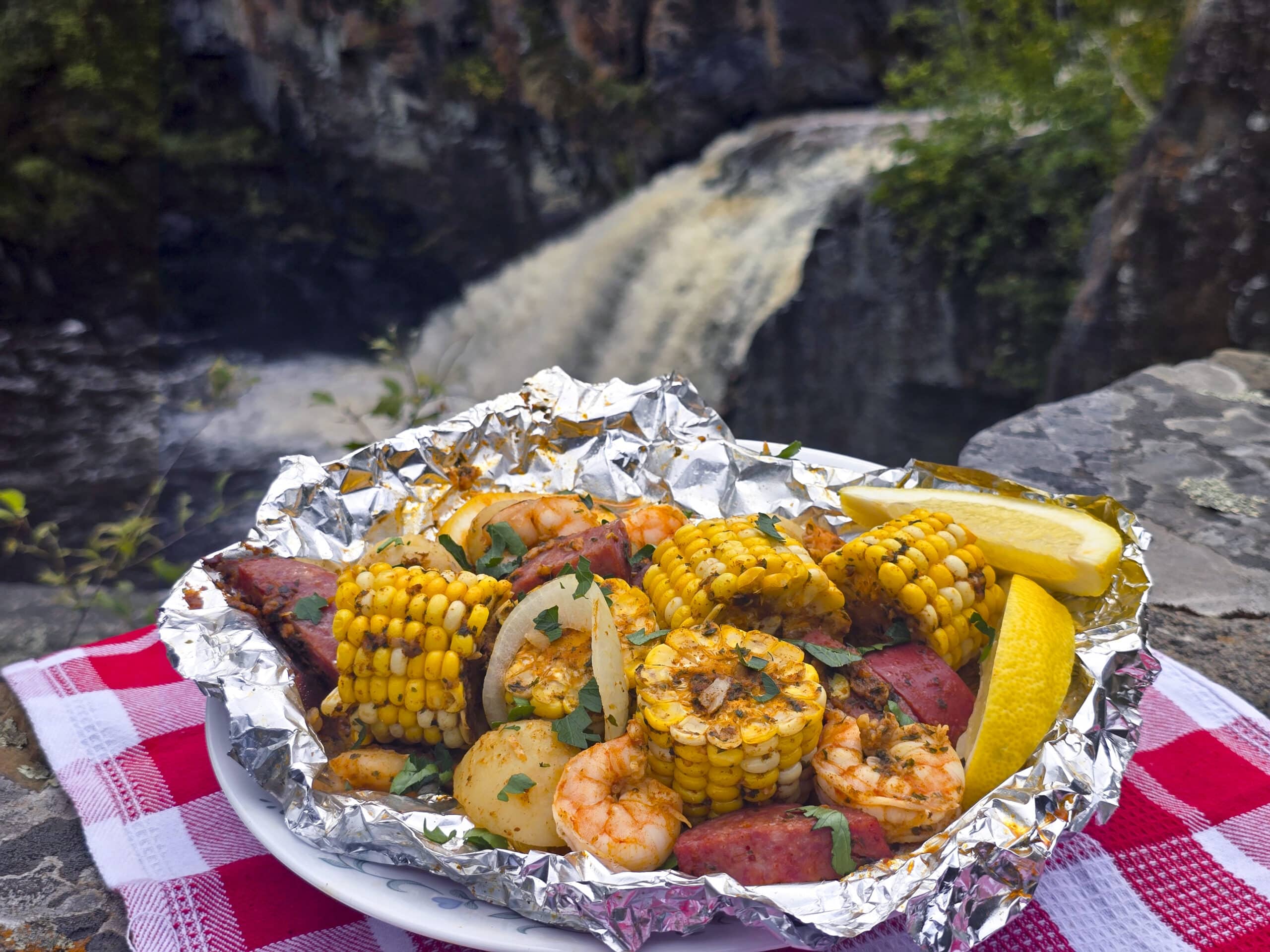 A large serving of seafood boil foil packet meal, on a plate. It has chunks of corn, sausage, potatoes, and shrimp in an obviously heavily seasoned sauce.