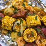 A large serving of shrimp boil foil packet meal, on a plate. It has chunks of corn, sausage, potatoes, and shrimp in an obviously heavily seasoned sauce.