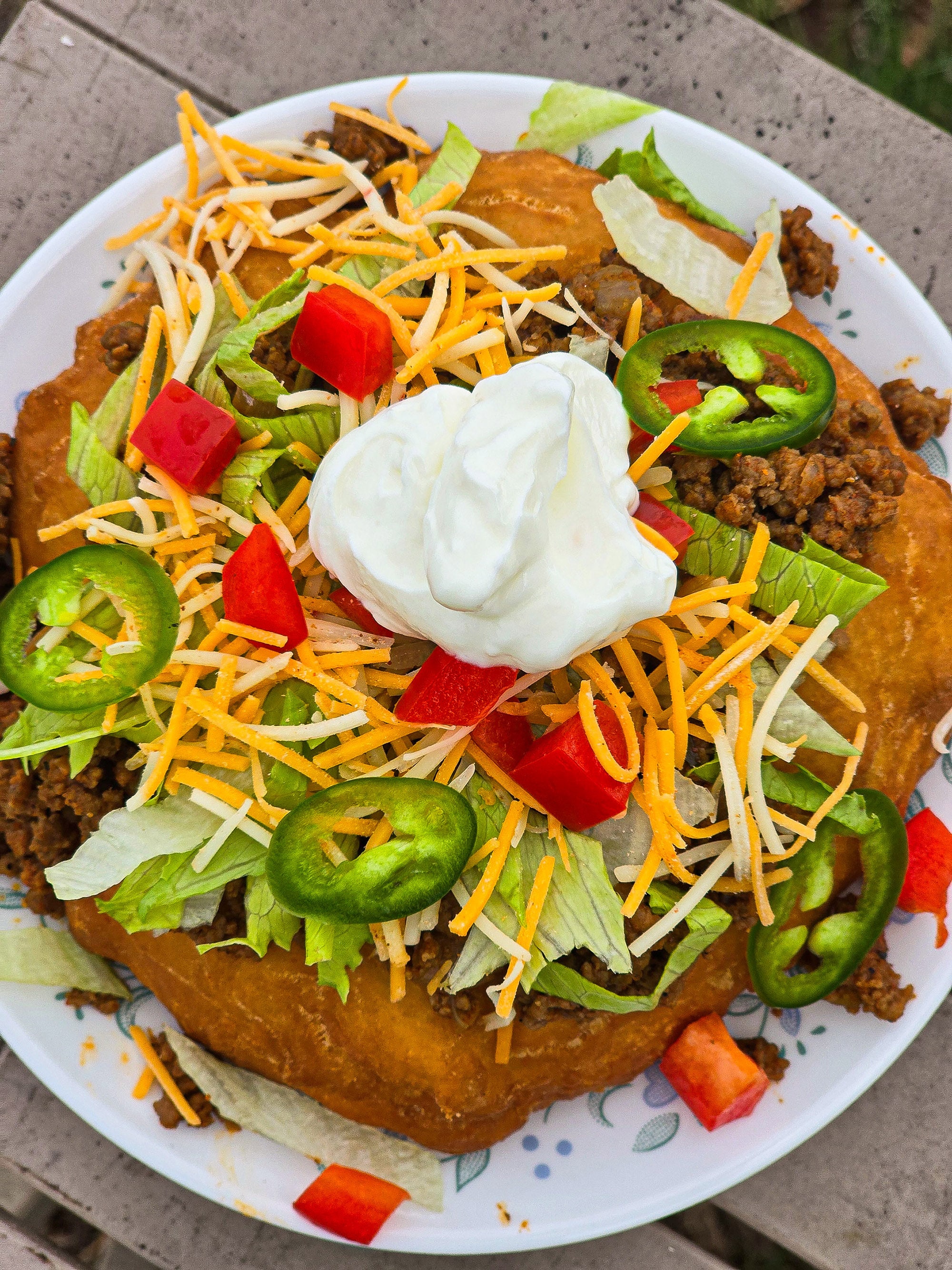 And Indian Taco made from a piece of fry bread, seasoned ground meat taco filling, lettuce, chopped red peppers, cheese, sour cream, and jalapeno pepper slices.